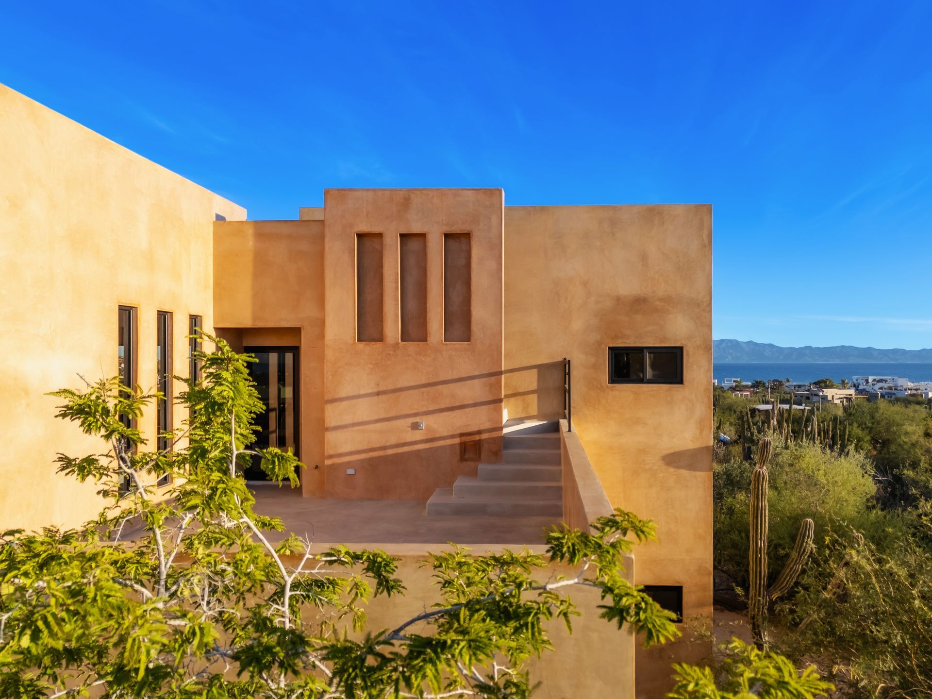 A multi-level, desert-style house with tan stucco walls, clean geometric lines, a stone staircase, and distant water.