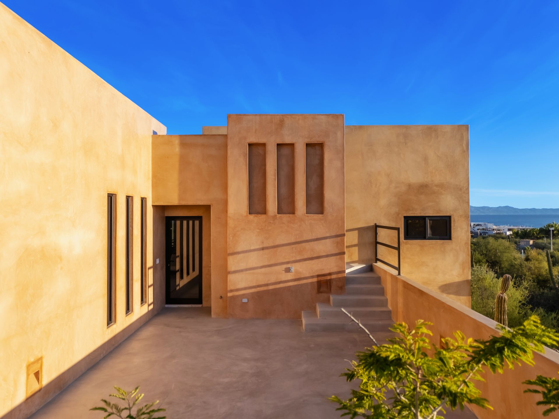 Modern stucco building exterior with a tan finish, narrow vertical windows, a doorway, and steps under a blue sky.