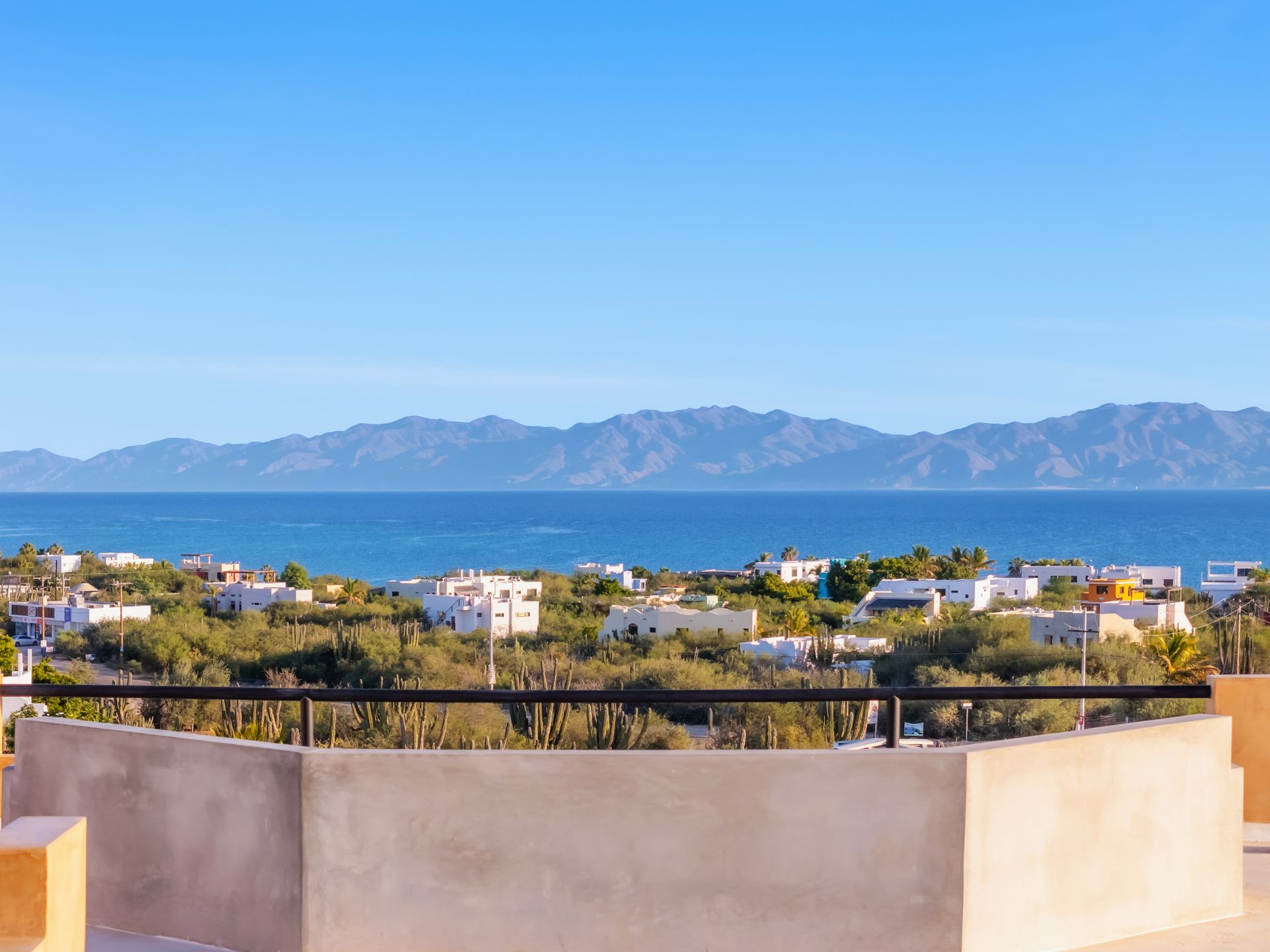 A scenic view of white buildings nestled in lush greenery, overlooking a blue ocean with distant mountains under a clear sky.