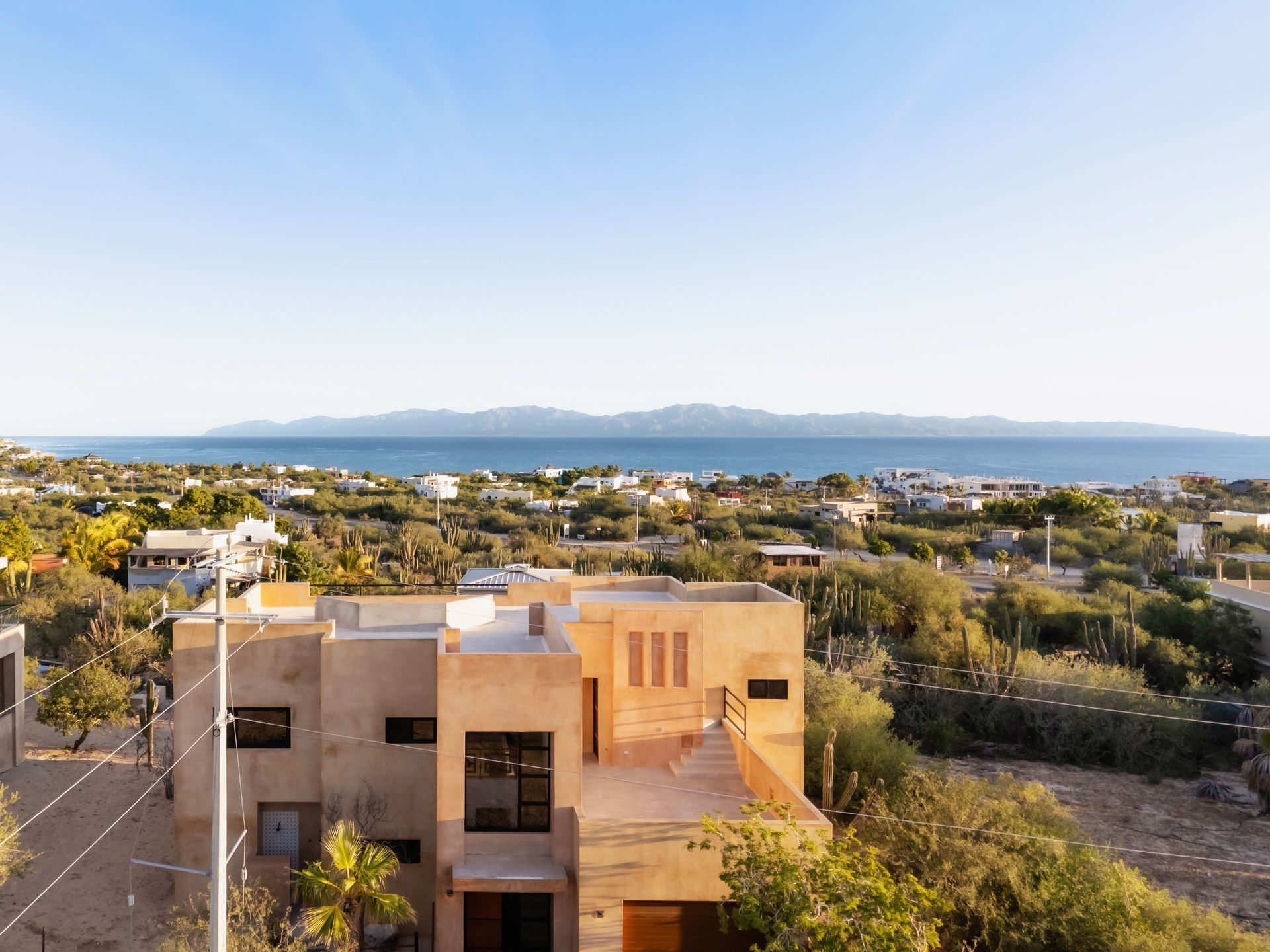A tan, modern multi-story house overlooking a vast coastal landscape with the ocean and distant mountains in the background.