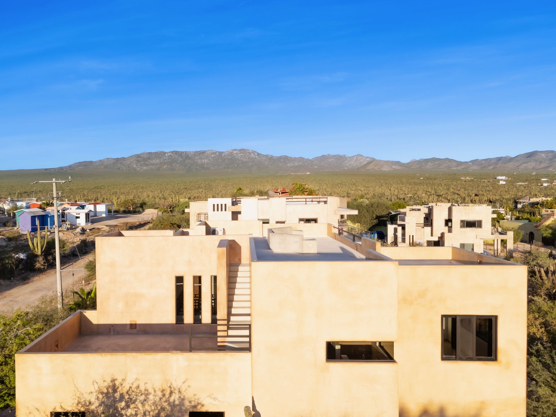 A modern, tan-colored minimalist villa with flat rooftops, set against a desert landscape with mountains in the background.