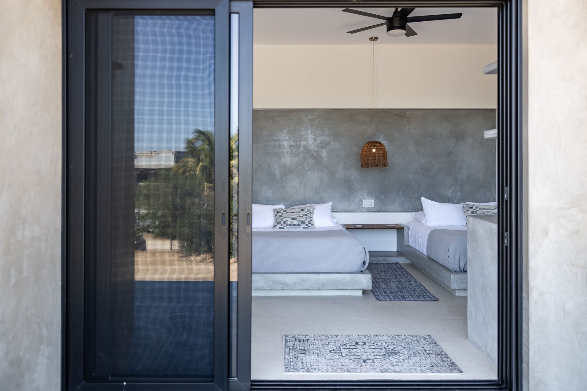 A view through a sliding glass door into a modern bedroom with two queen beds, concrete walls, and a ceiling fan.