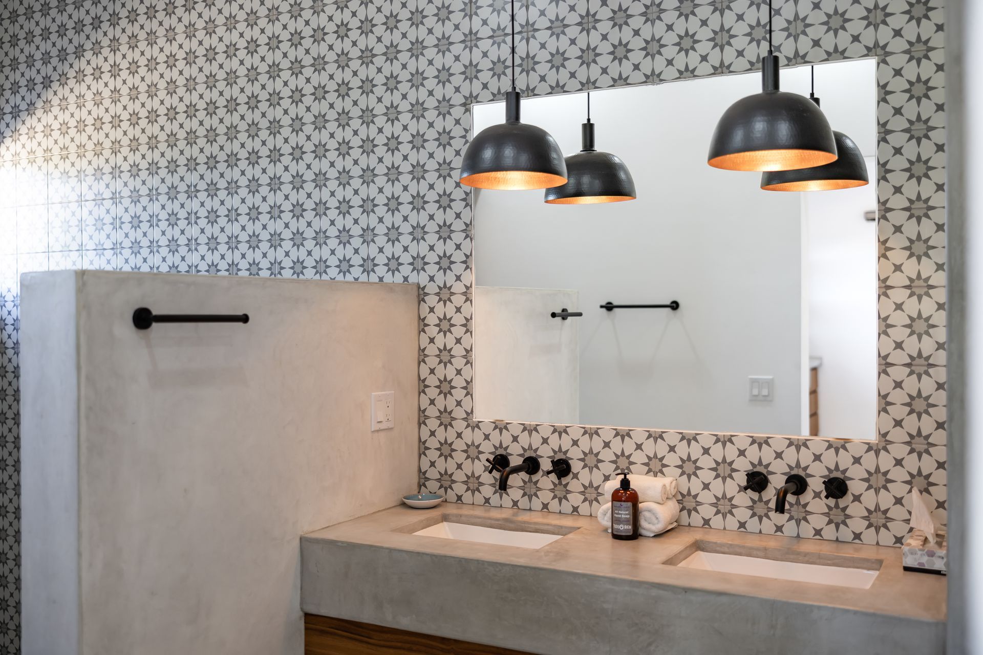 A bathroom vanity featuring a double concrete sink, patterned tile wall, mirror, and three hanging black dome lights.