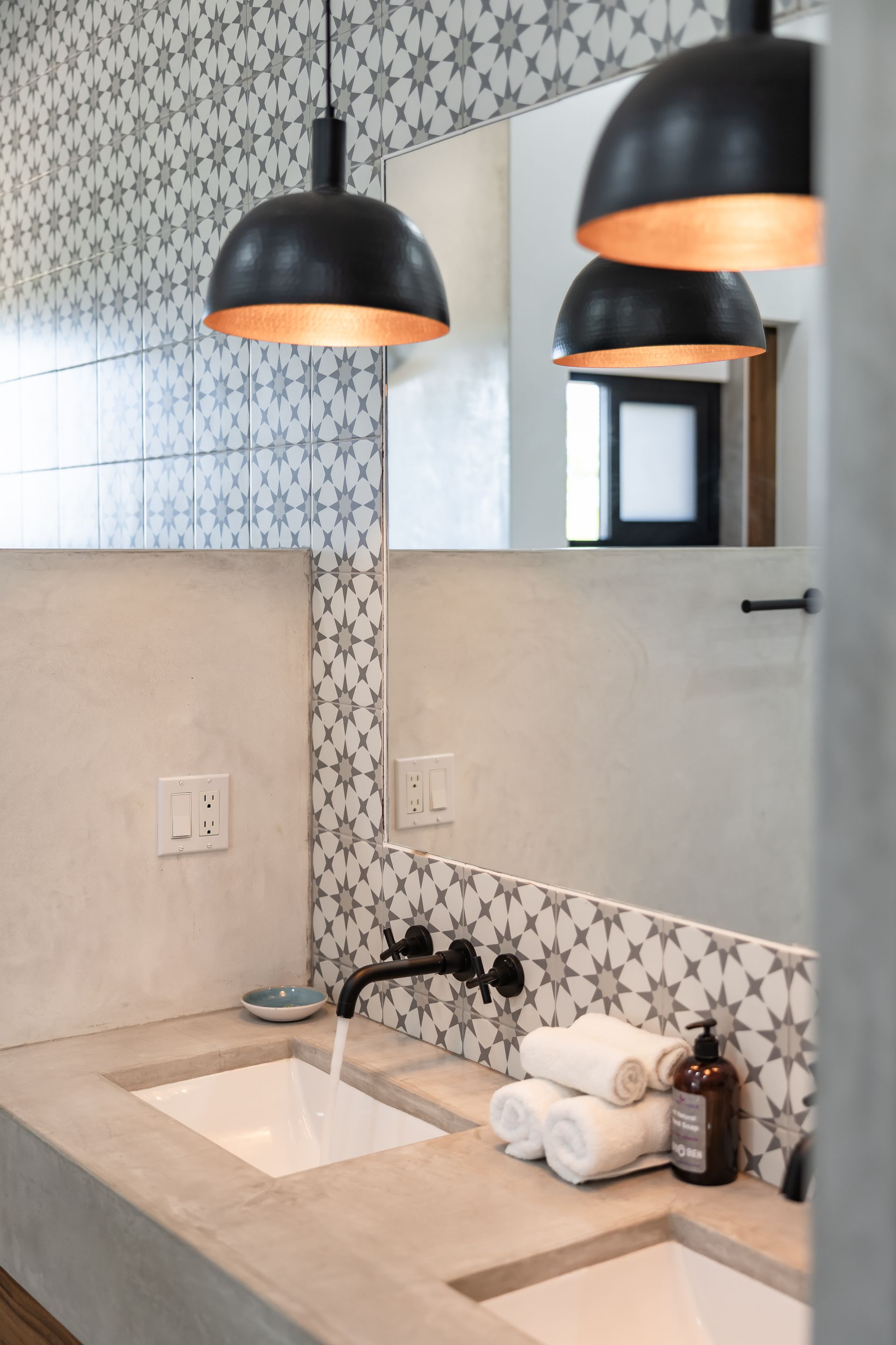 Double concrete vanity with white vessel sinks, wall-mounted faucets, and black pendant lights against patterned tiles.