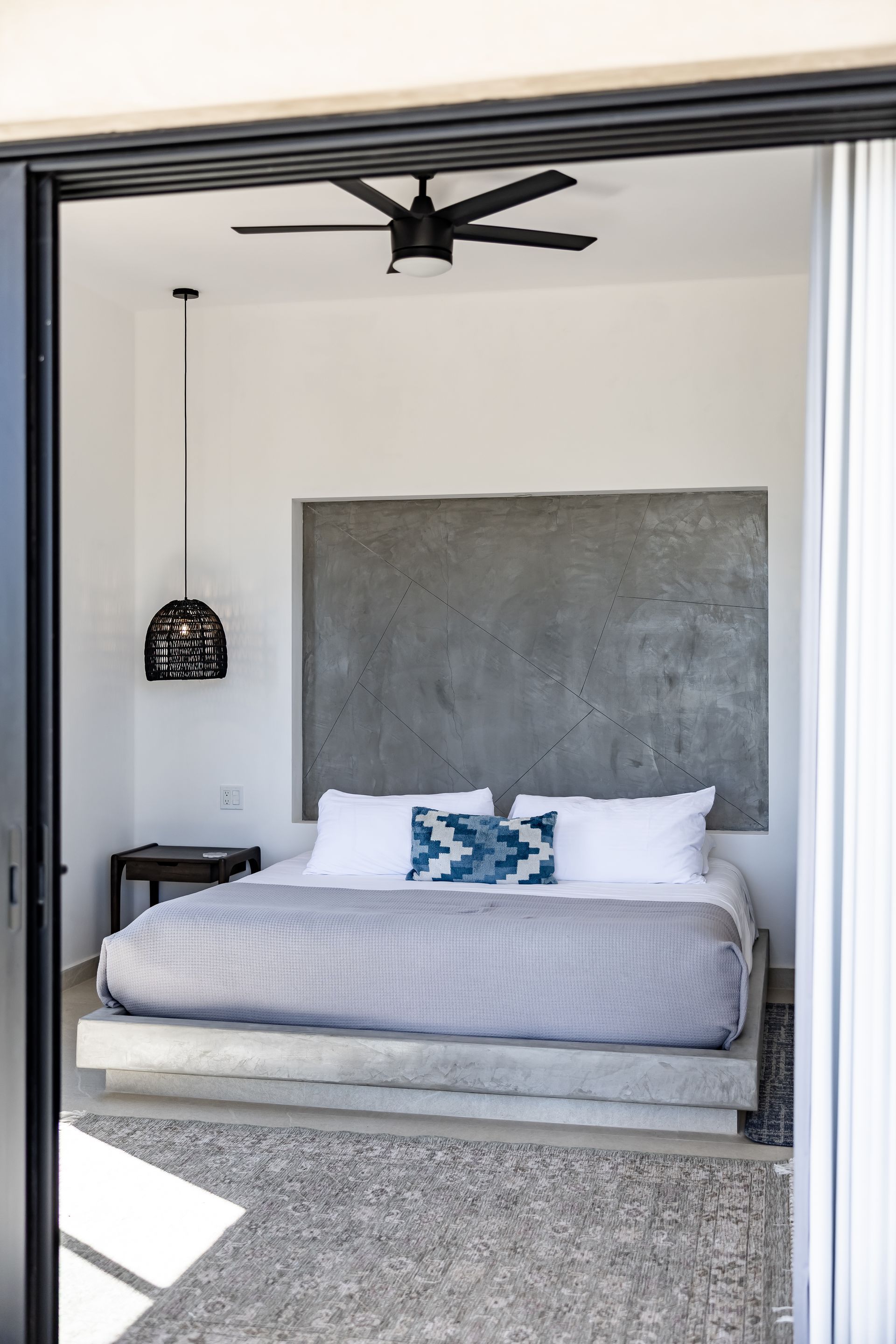 A minimalist bedroom featuring a bed with white linens on a concrete base, a grey textured headboard, and hanging lamp.