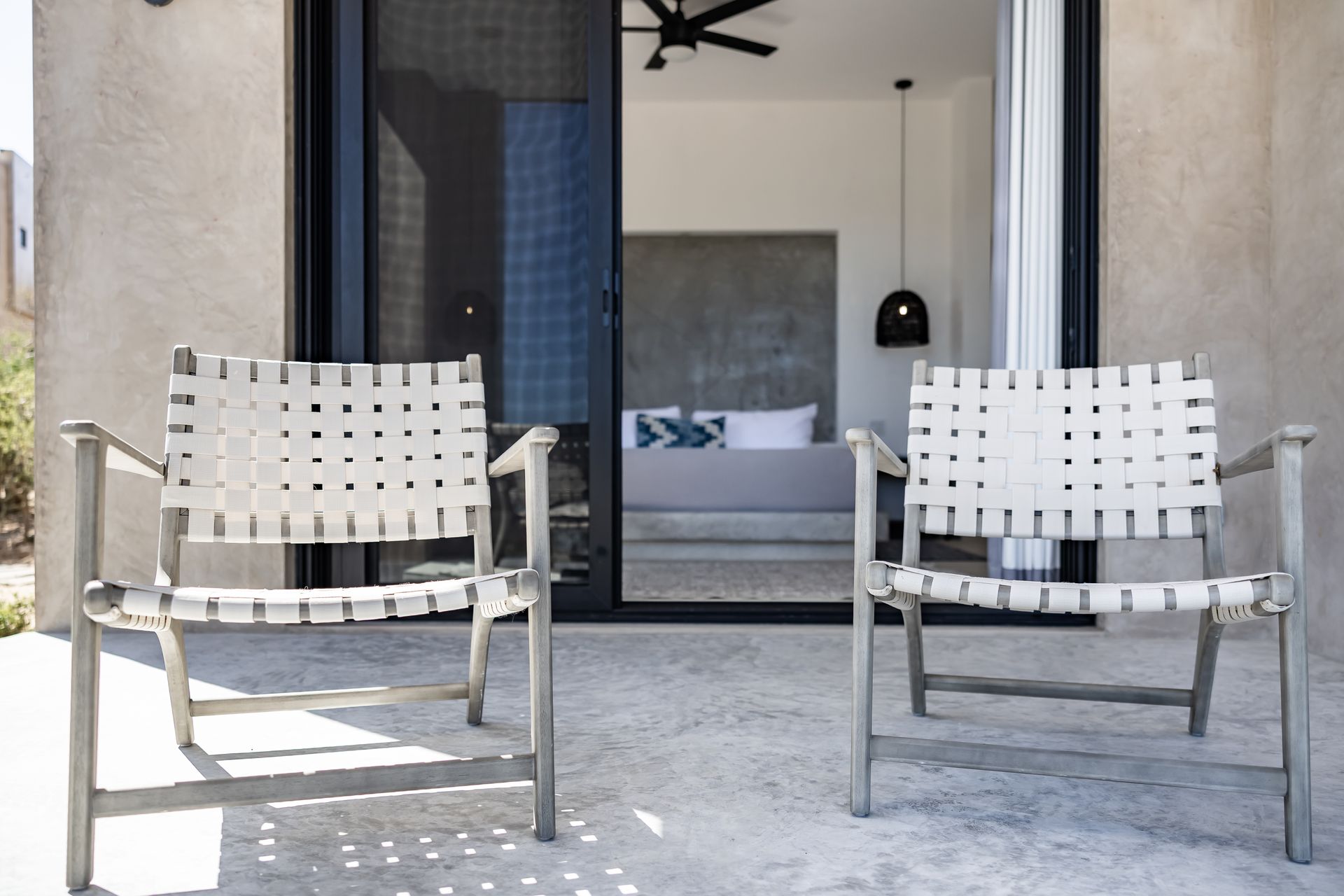 Two woven armchairs sit on a concrete patio in front of a sliding glass door opening into a bedroom.