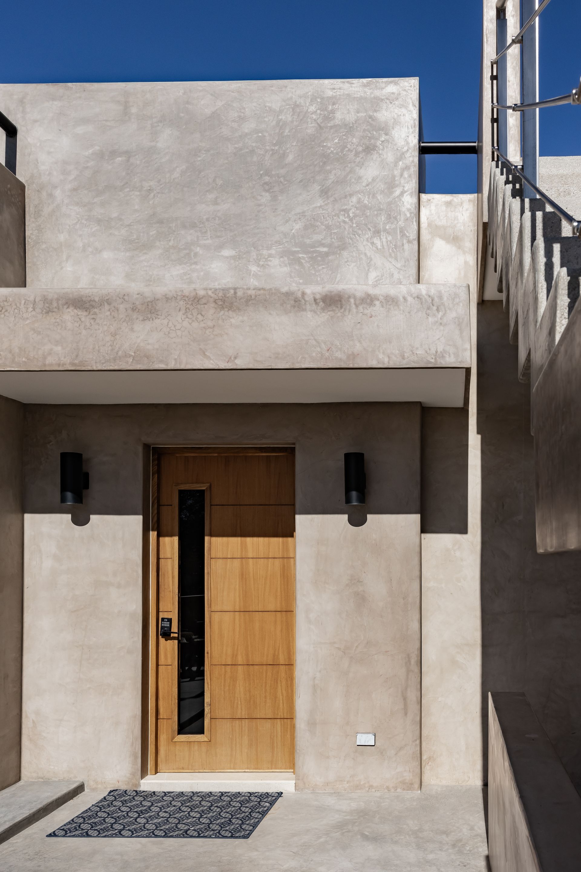 A modern home entrance with a light wood door featuring a vertical glass panel, flanked by two black wall lights in daylight.