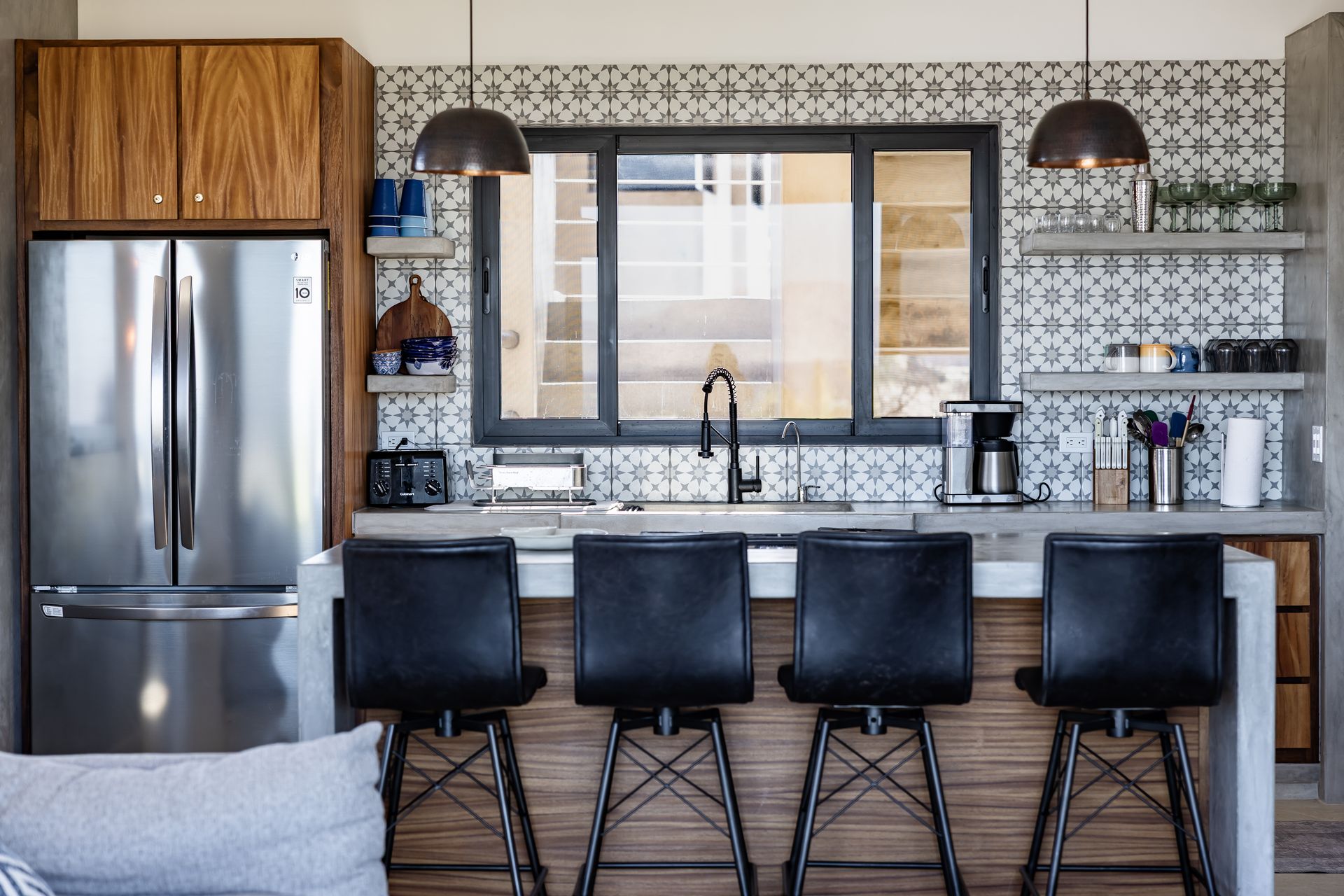 Modern kitchen with four black bar stools at a concrete island, a stainless refrigerator, and patterned tile backsplash.