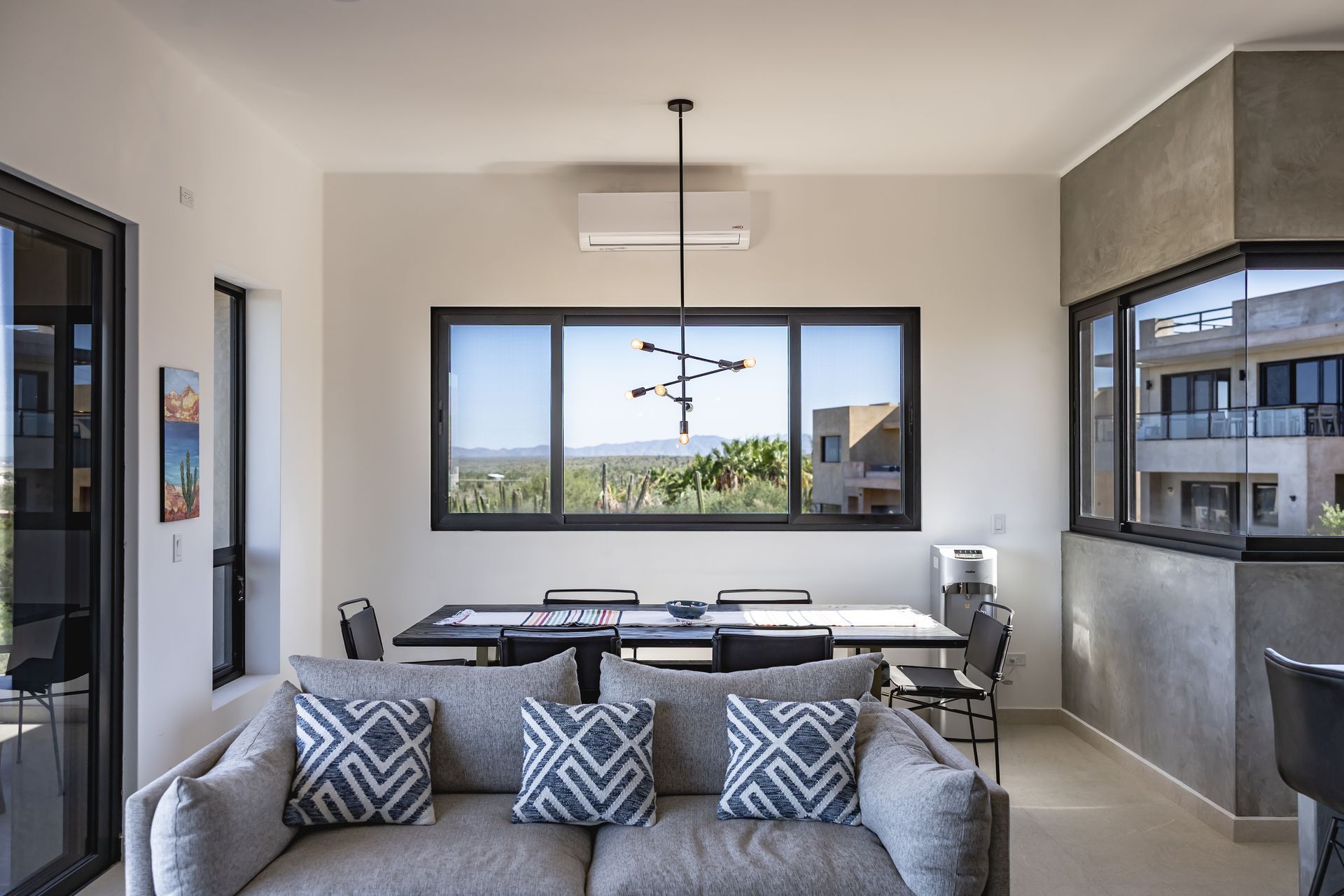 A modern living and dining room with a grey sofa in the foreground, a dining set, and a view of a landscape through windows.