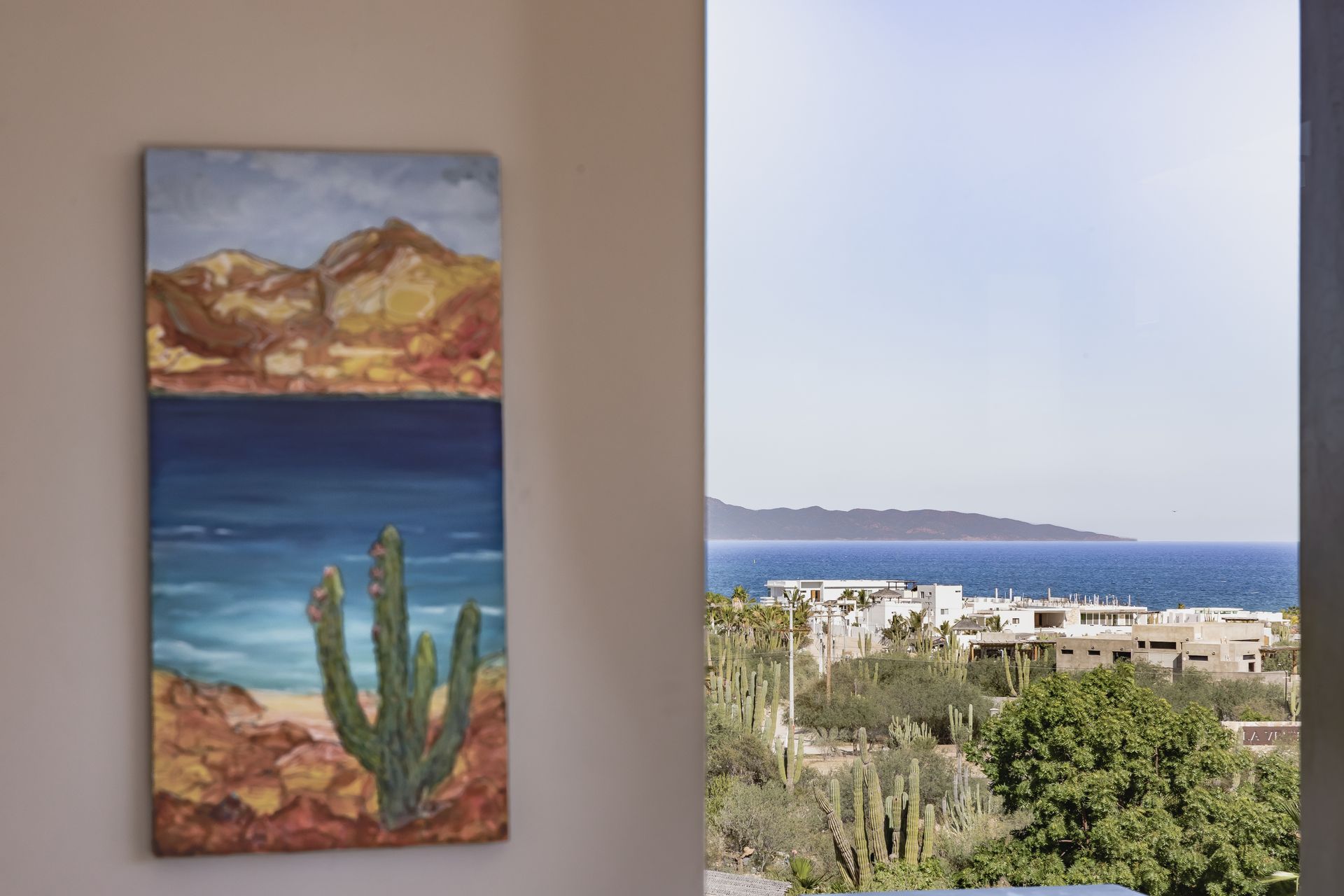 A painting of a cactus on a beach hangs on a wall next to a window overlooking a coastal landscape with trees and buildings.