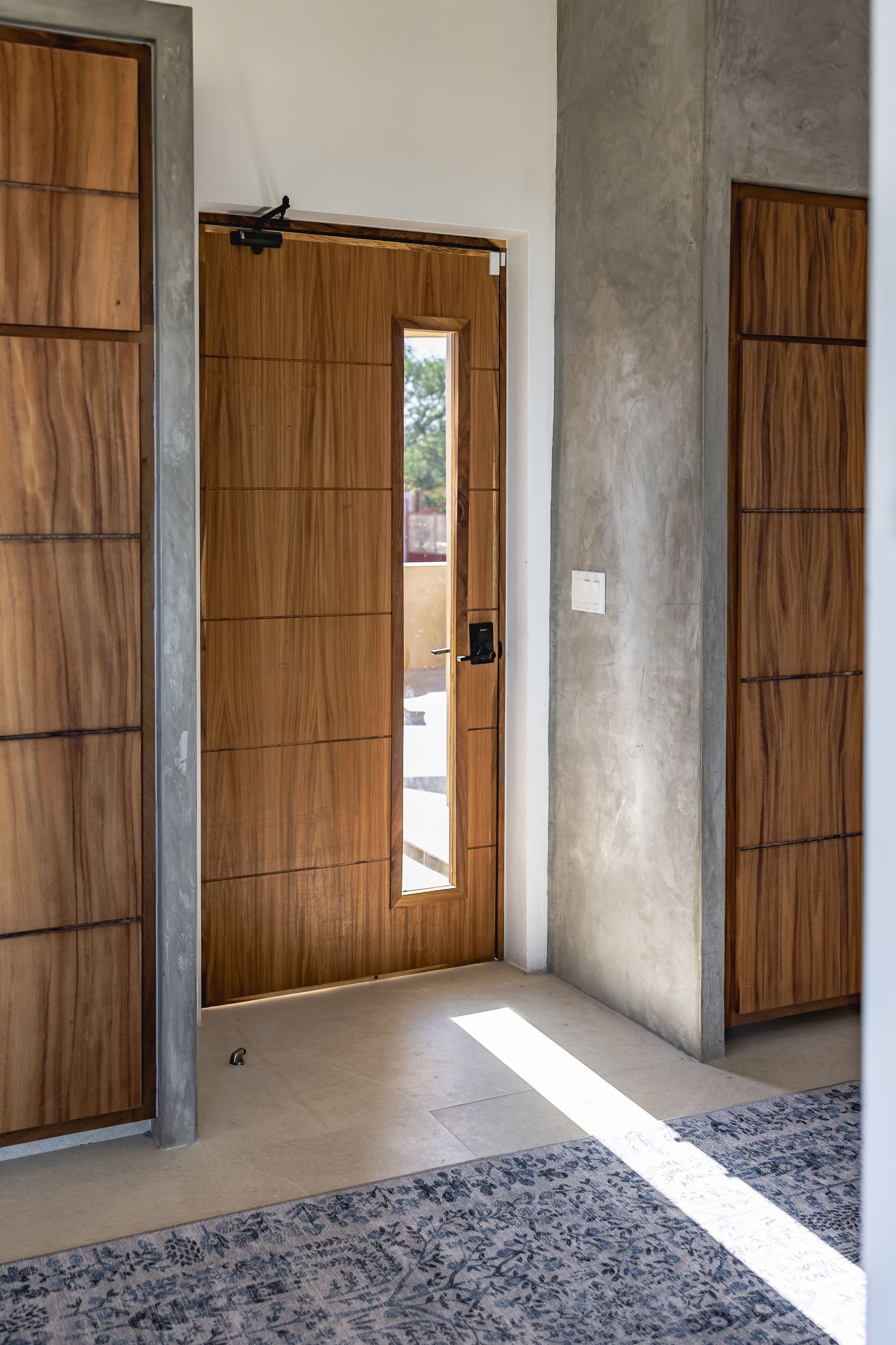 A modern entryway with wooden panel doors, concrete walls, a vertical window, and a patterned rug on the floor.