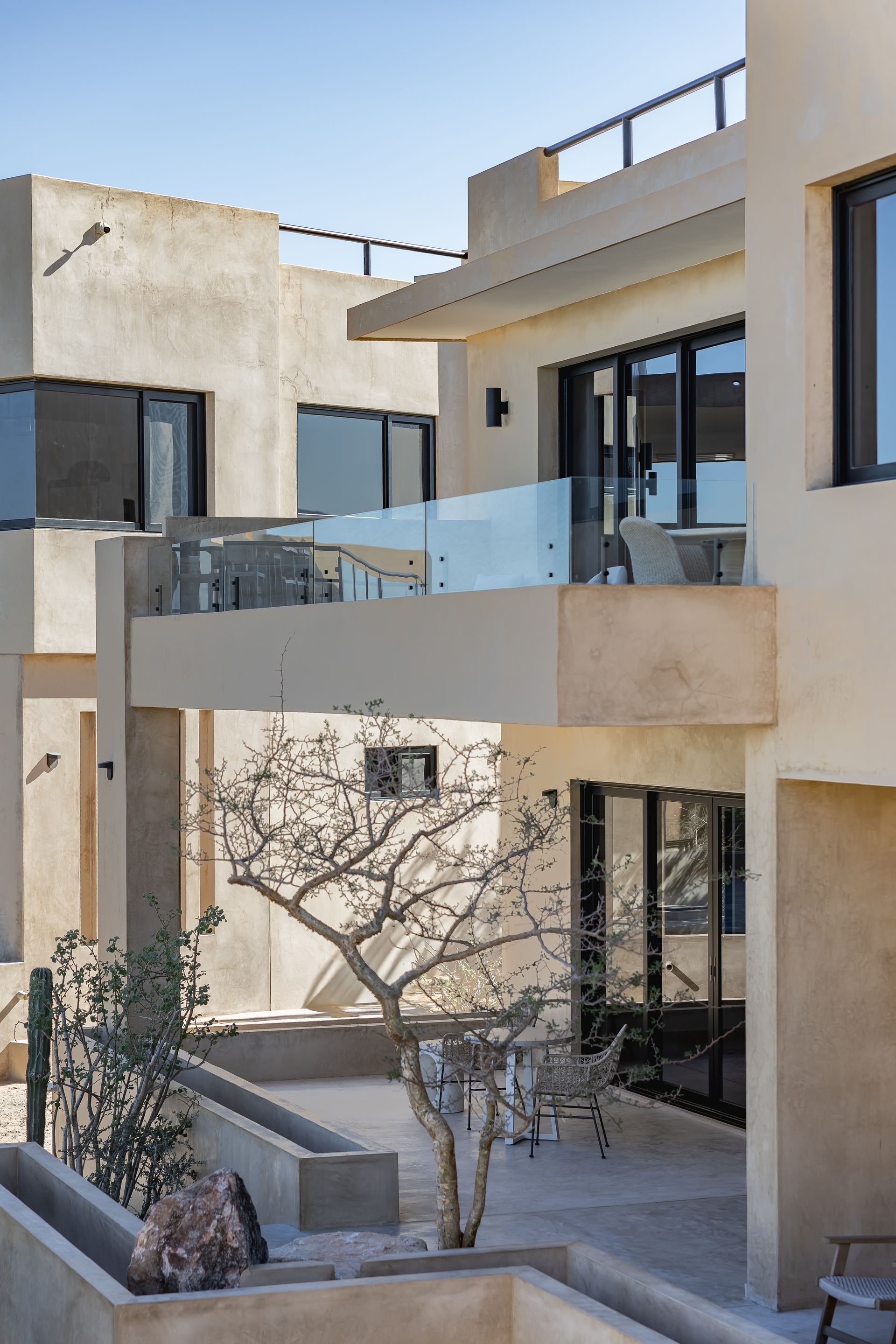 Modern, beige multi-level villas with balconies and a patio area featuring a bare tree and stone planters.