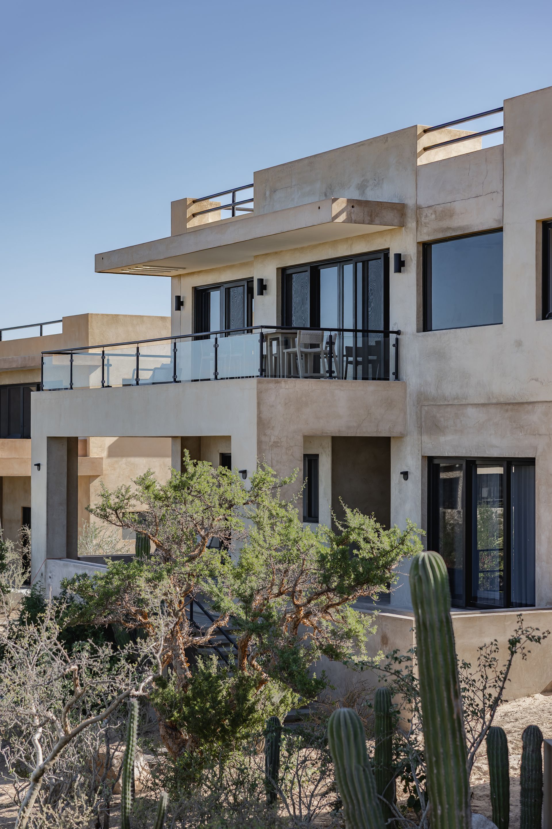 A modern, multi-story tan stucco home with balconies and dark-framed windows, surrounded by desert cacti and shrubs.
