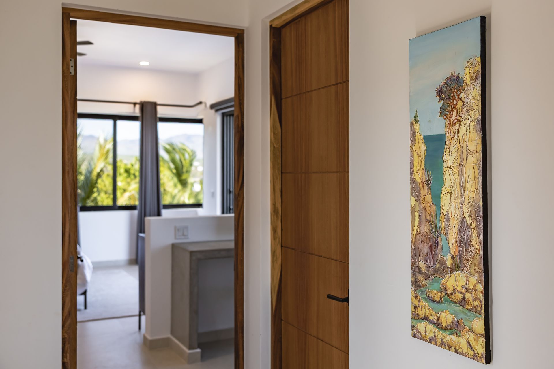 A hallway with a wooden door, a tall, colorful landscape painting on the wall, and a view into a bright bedroom.