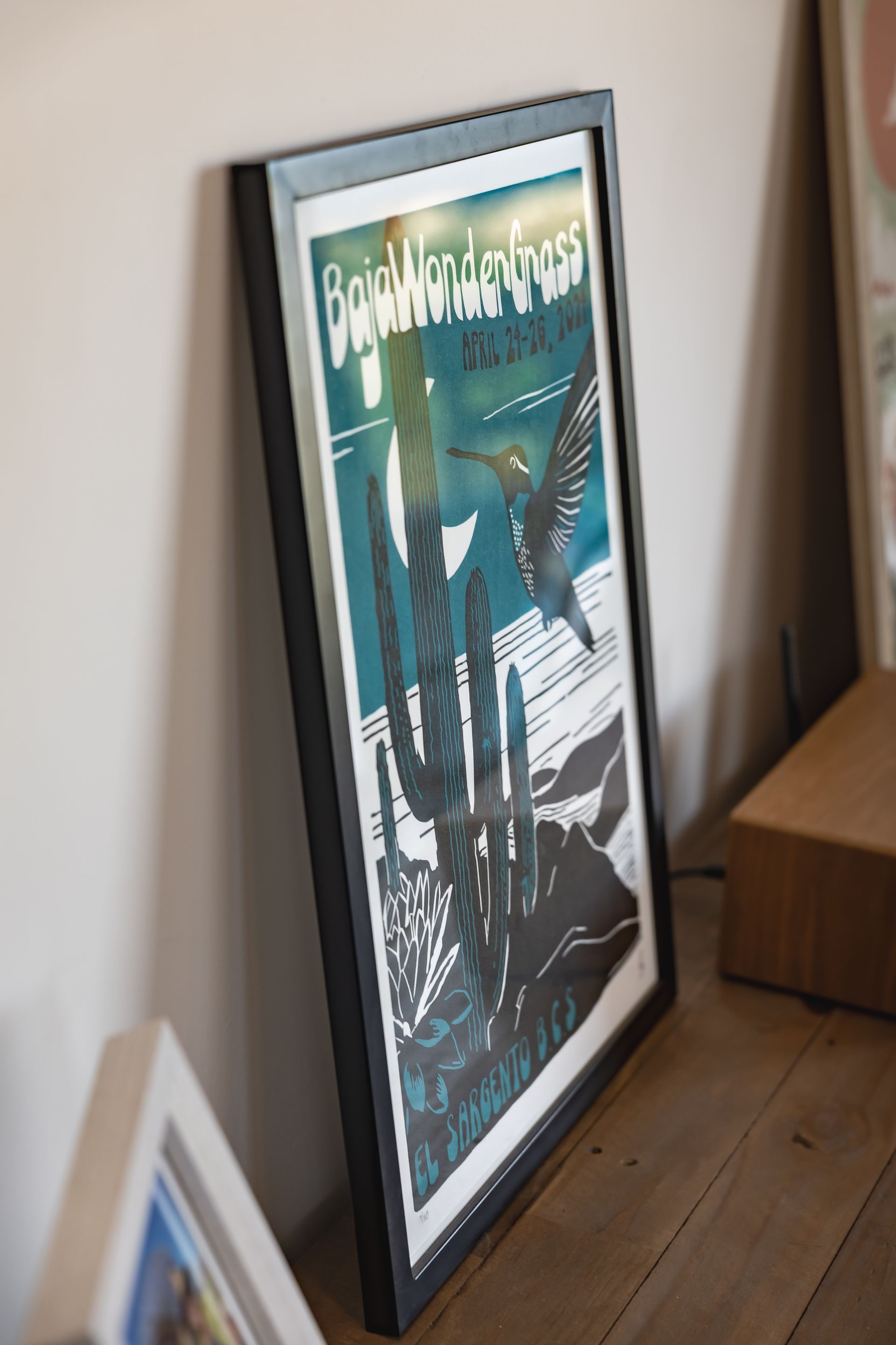A framed desert-themed poster featuring a hummingbird and a saguaro cactus, resting on a wooden surface.