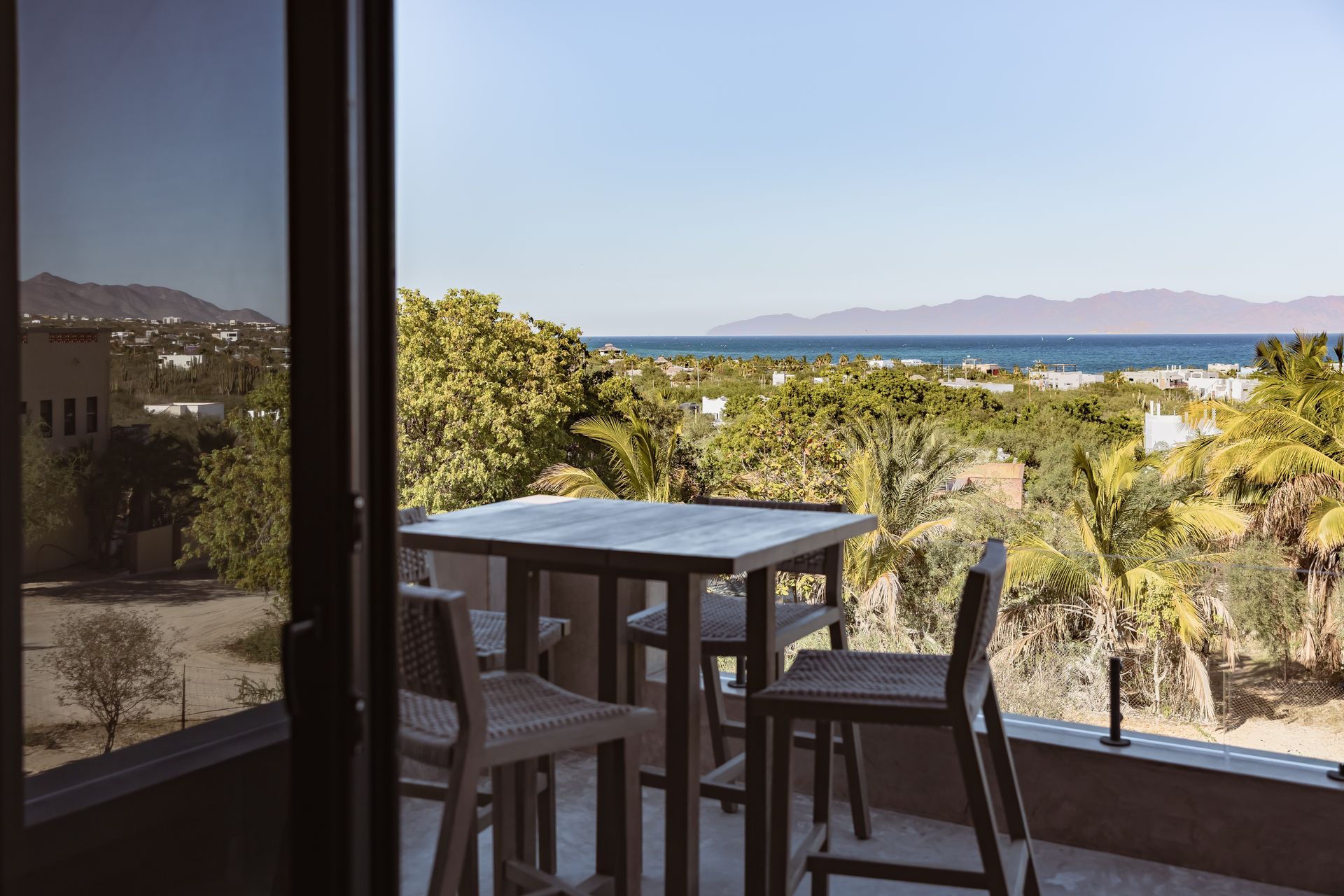 A square patio table and chairs overlook a lush landscape, coastline, and distant mountains under a clear blue sky.