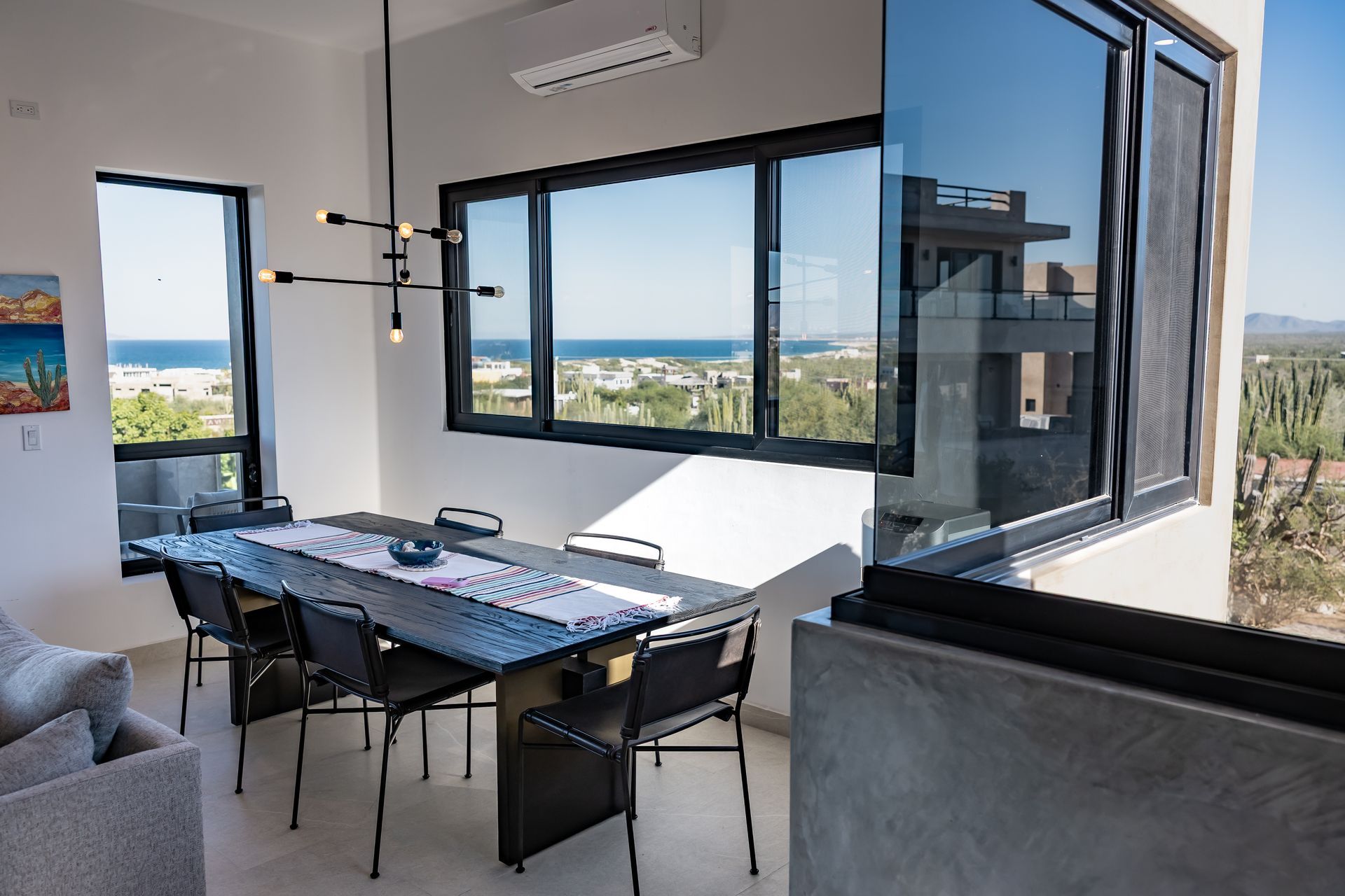 A dining room with a long black table and chairs overlooking a scenic coastal view through large floor-to-ceiling windows.