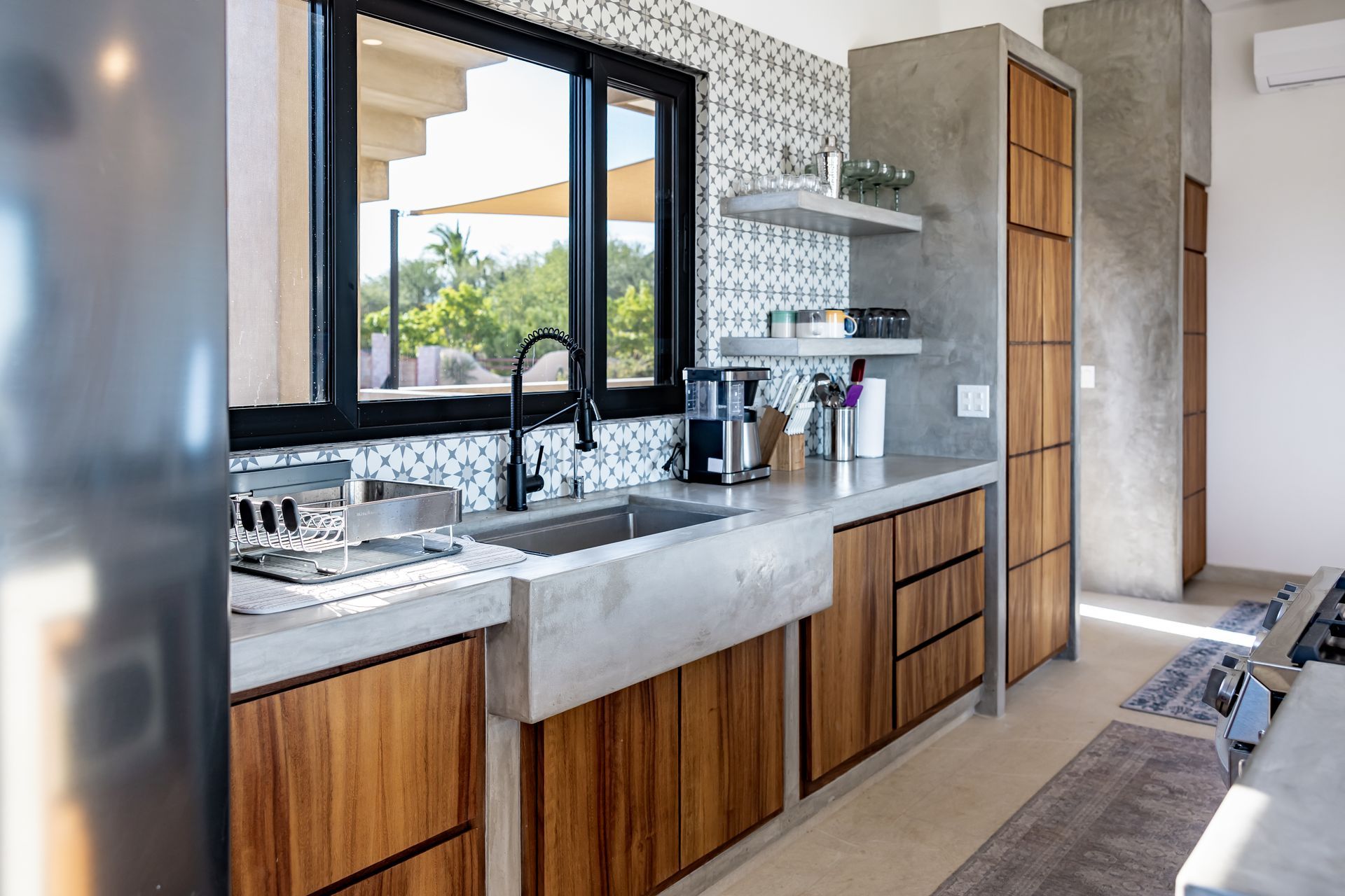 A modern kitchen featuring wood cabinets, concrete countertops, a farmhouse sink, and a patterned tile backsplash.