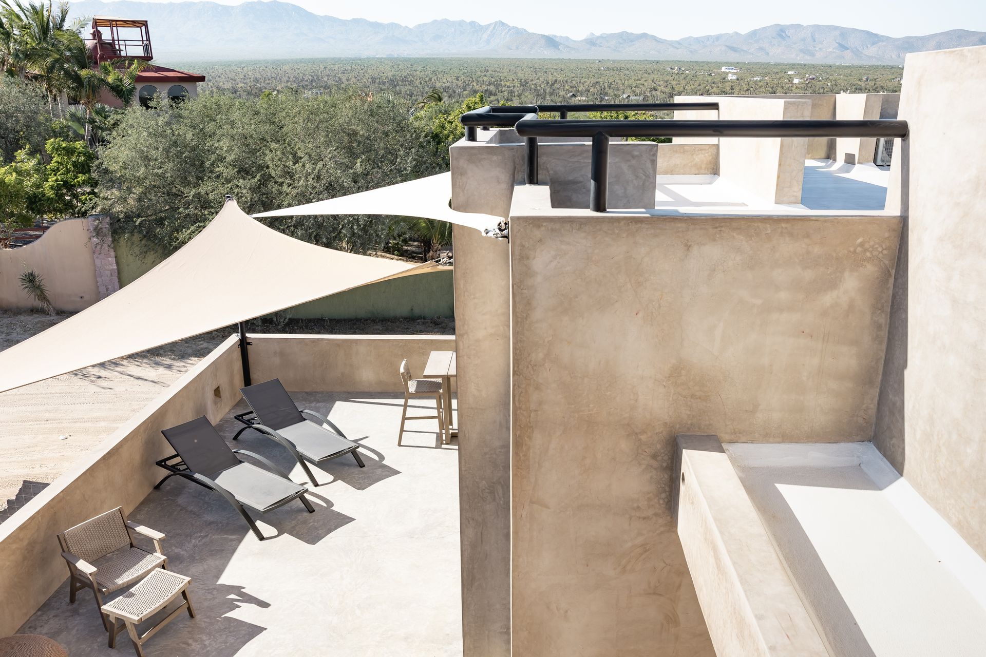 A sunny rooftop terrace featuring lounge chairs, a table, and a beige sun sail, overlooking a desert landscape.