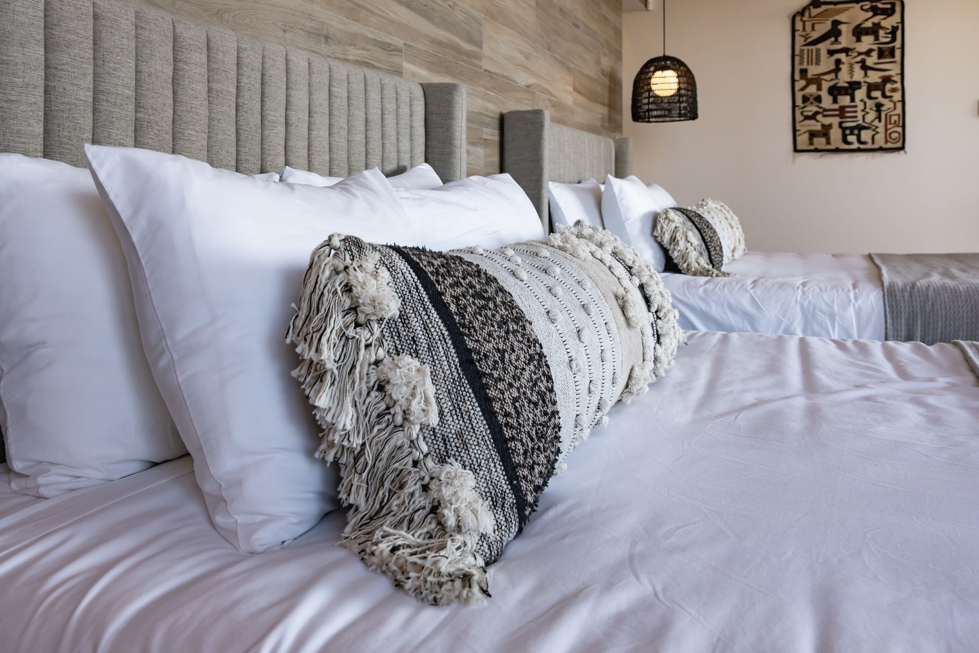 A bedroom with two beds featuring white linens, gray channeled headboards, and a patterned, fringed decorative pillow.