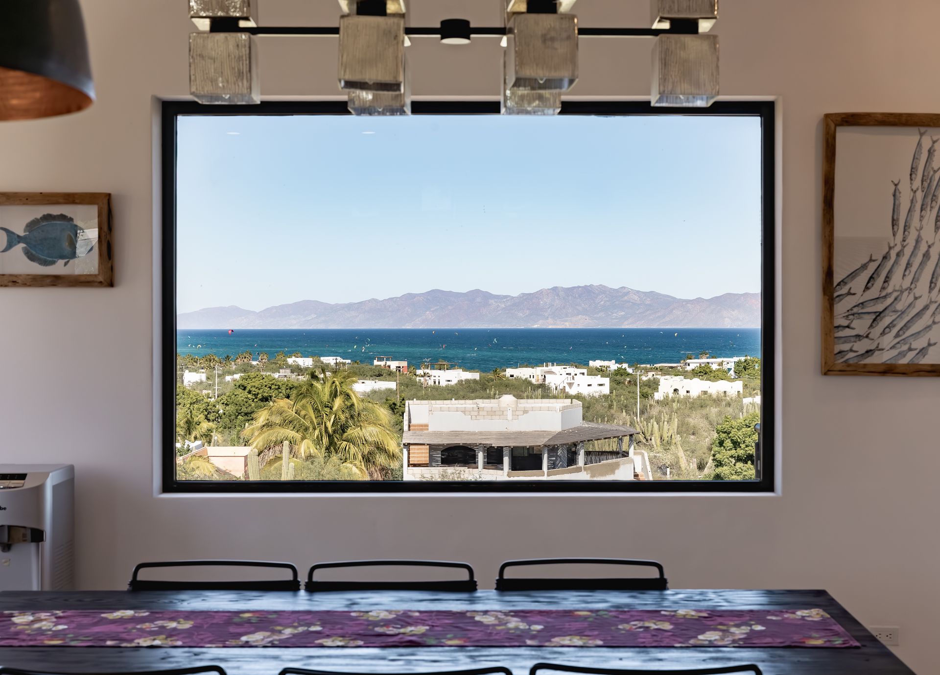A dining table with a purple runner faces a large window overlooking a lush landscape, the ocean, and distant mountains.