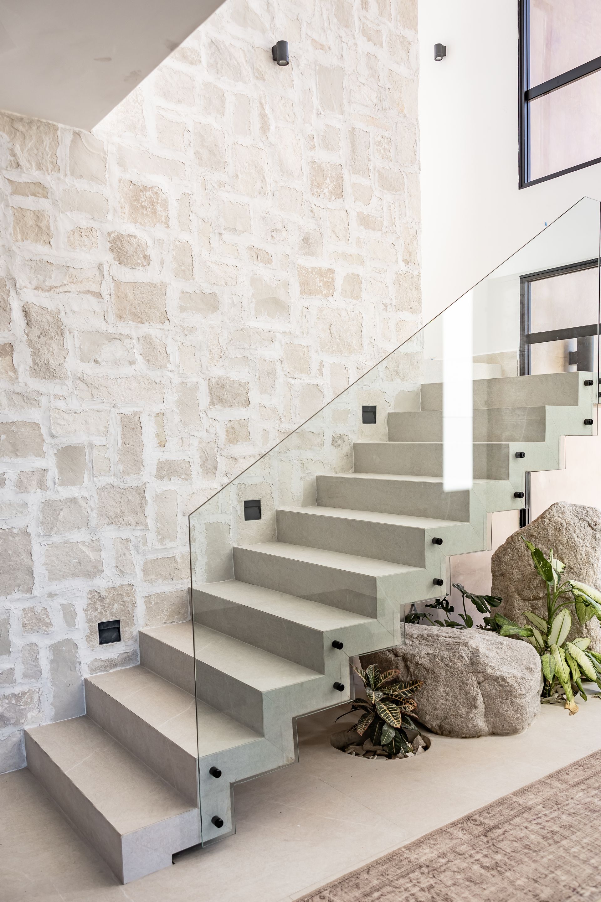 Modern staircase with concrete treads, a glass railing, and a stone accent wall, featuring natural rocks and indoor plants.