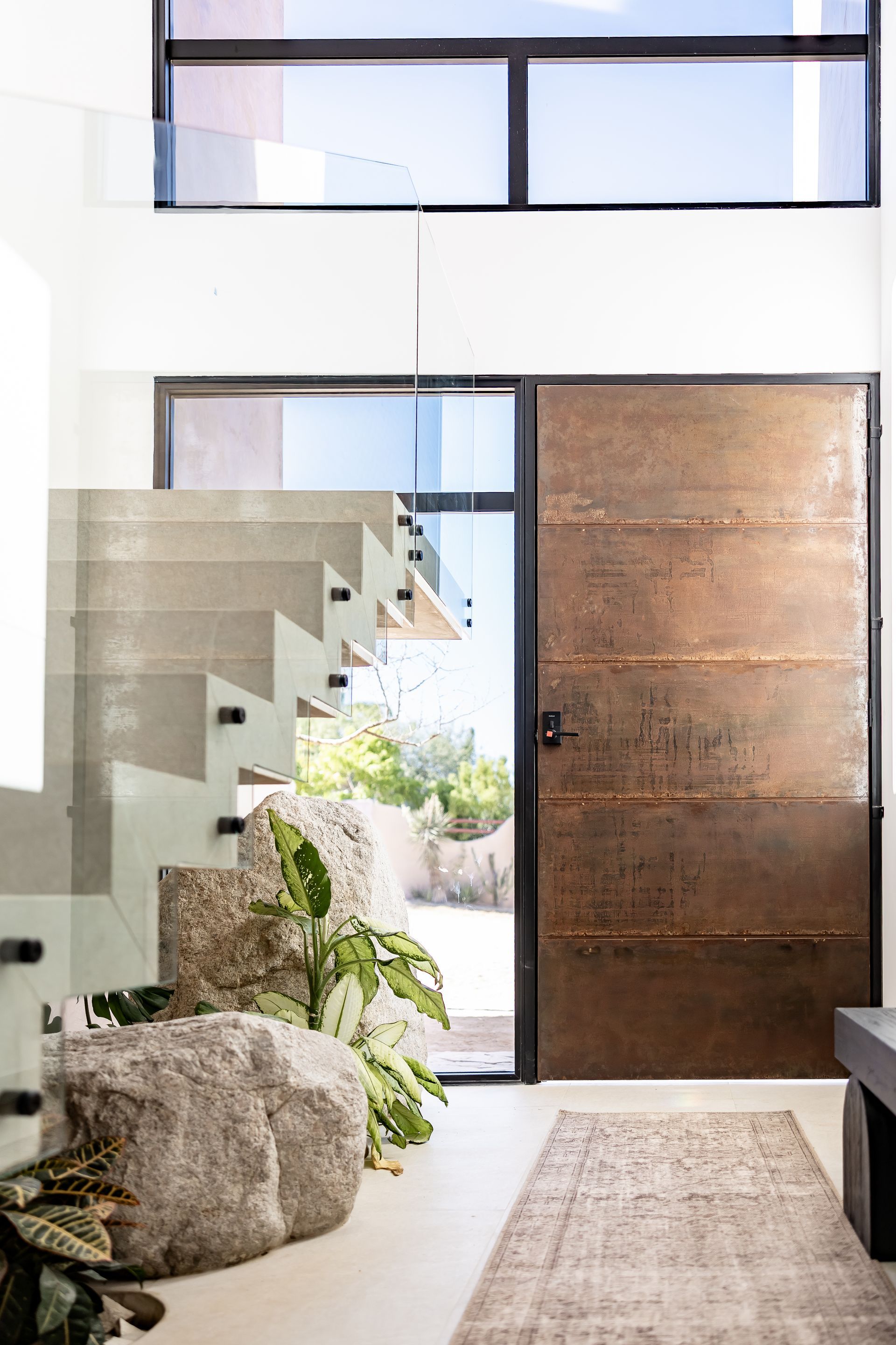 A modern entryway with a tall, rust-colored metal door, a floating concrete staircase, large boulders, and plants.