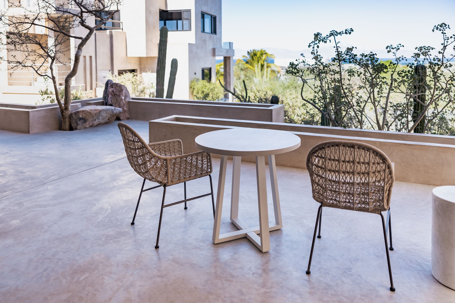 A small round table with two woven chairs on a sunny outdoor patio overlooking a desert landscape.