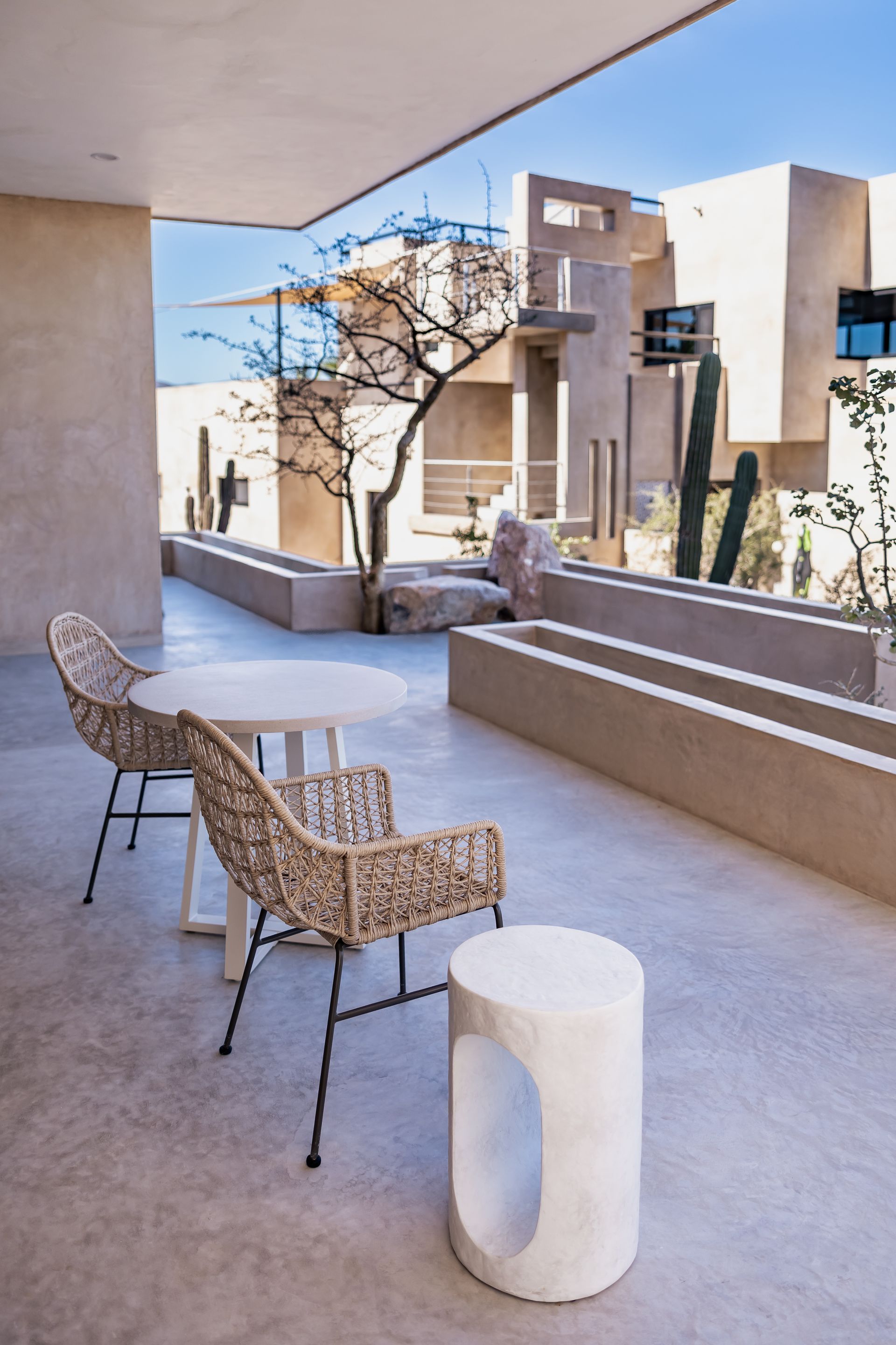 A sunlit patio with two woven chairs, a small table, a stone side table, and cacti against modern desert-style buildings.