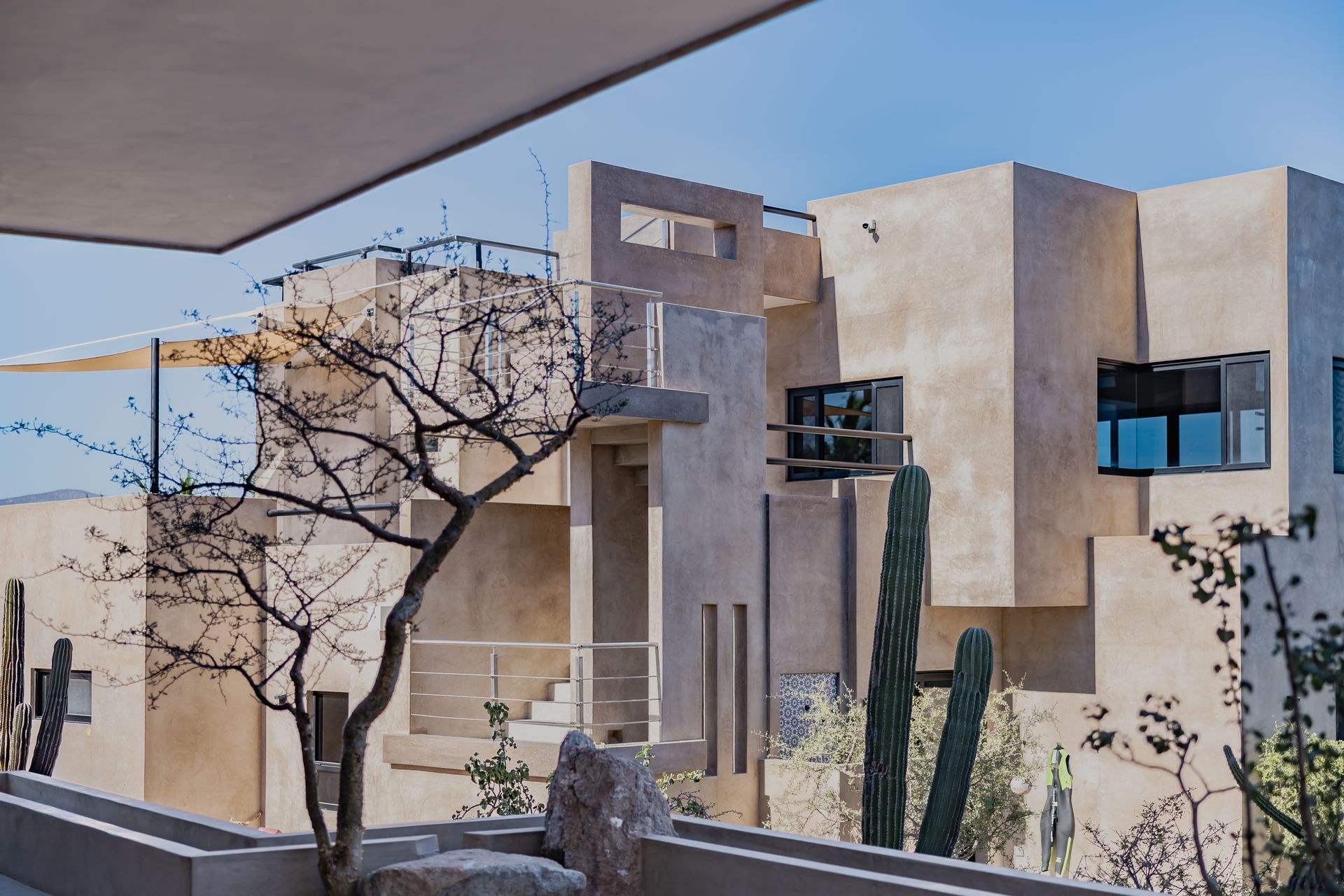 A tan, modern multi-level building with geometric architecture, surrounded by cacti and desert vegetation under a blue sky.