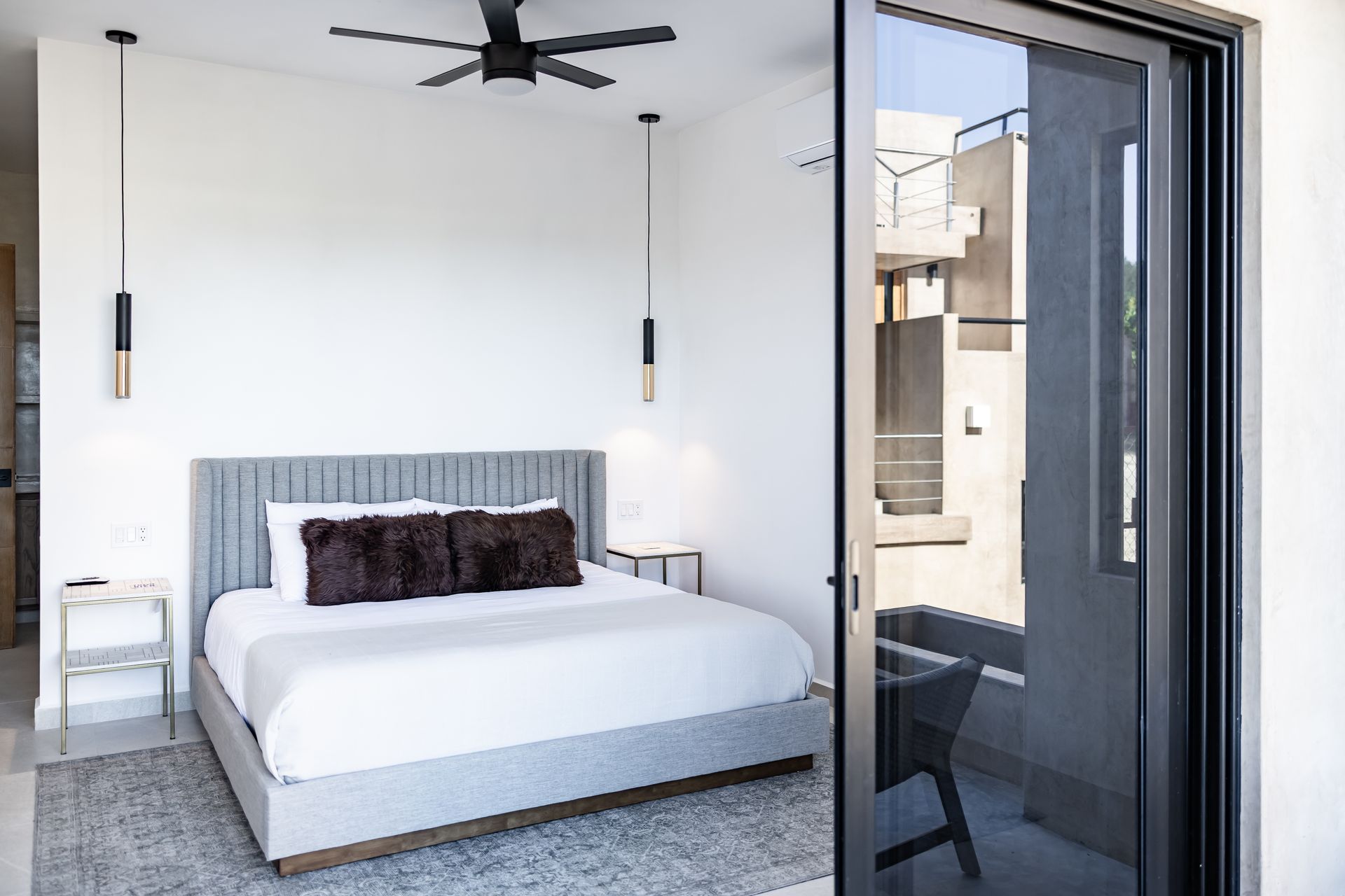 A modern, airy bedroom with a gray upholstered bed, white bedding, dark pillows, and a glass patio door.