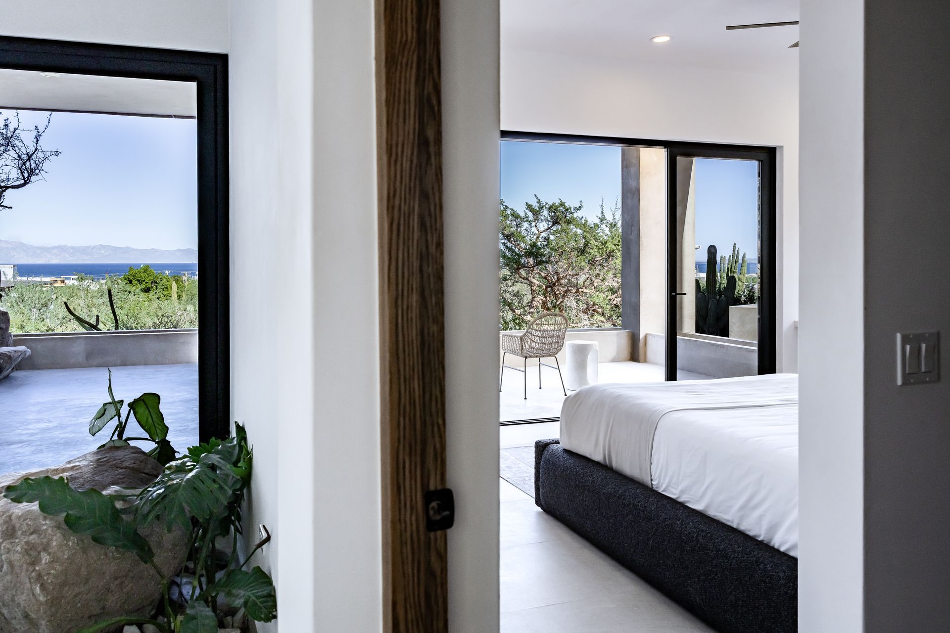 A view through a doorway into a bright bedroom with a bed and sliding doors opening to a scenic patio and ocean view.