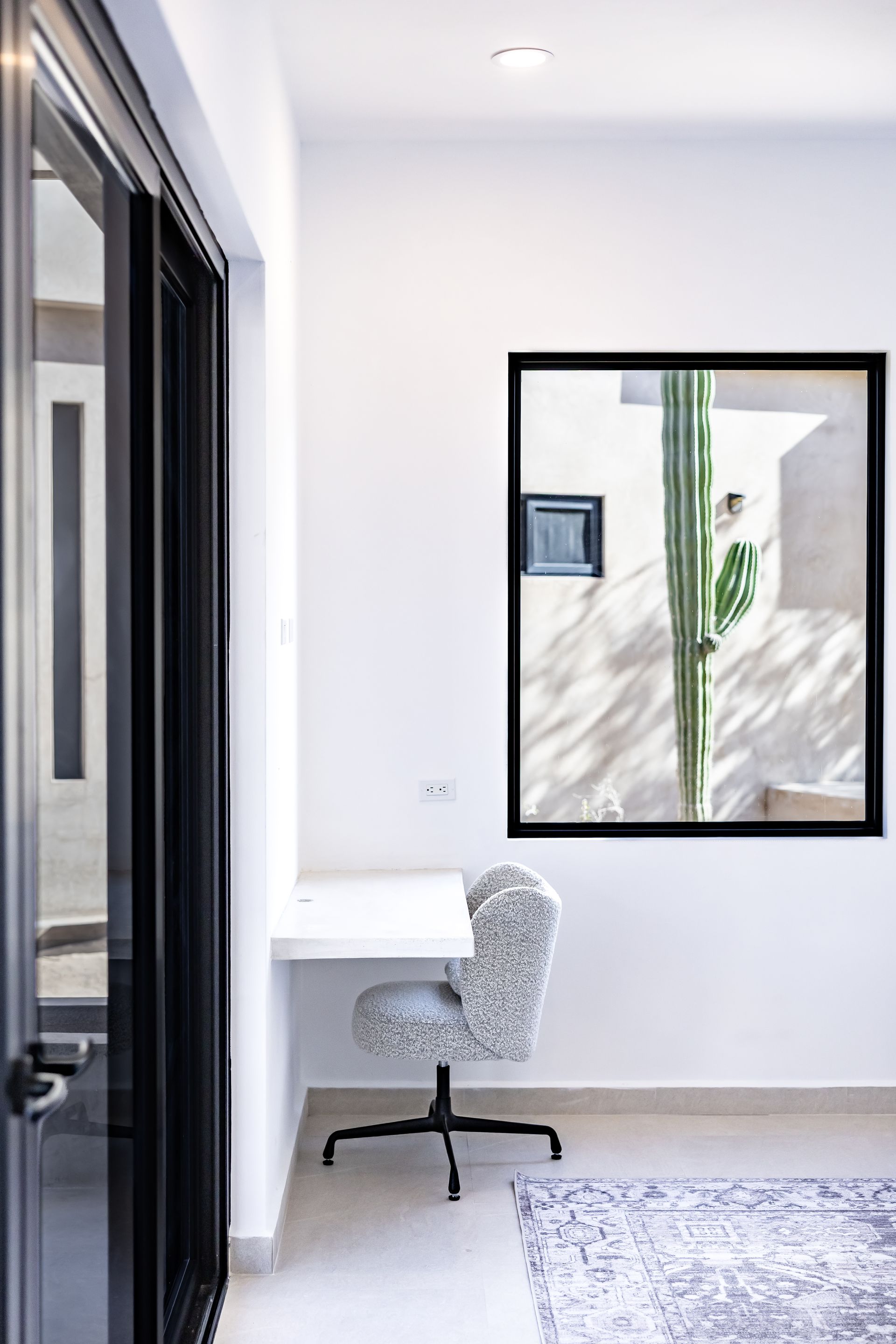 A modern, minimalist home office with a white floating desk, a patterned office chair, and a window view of a tall cactus.