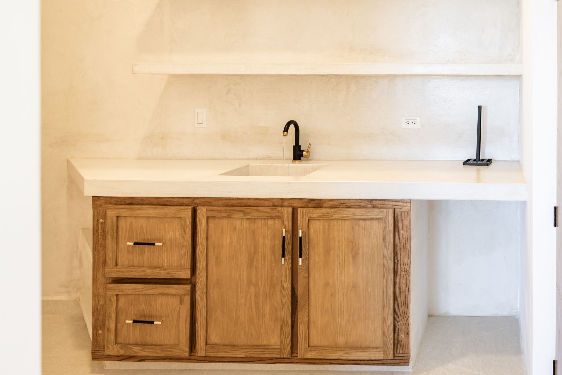 A minimalist kitchen vanity with a white countertop, sink, black faucet, and light oak wood cabinets set against white walls.