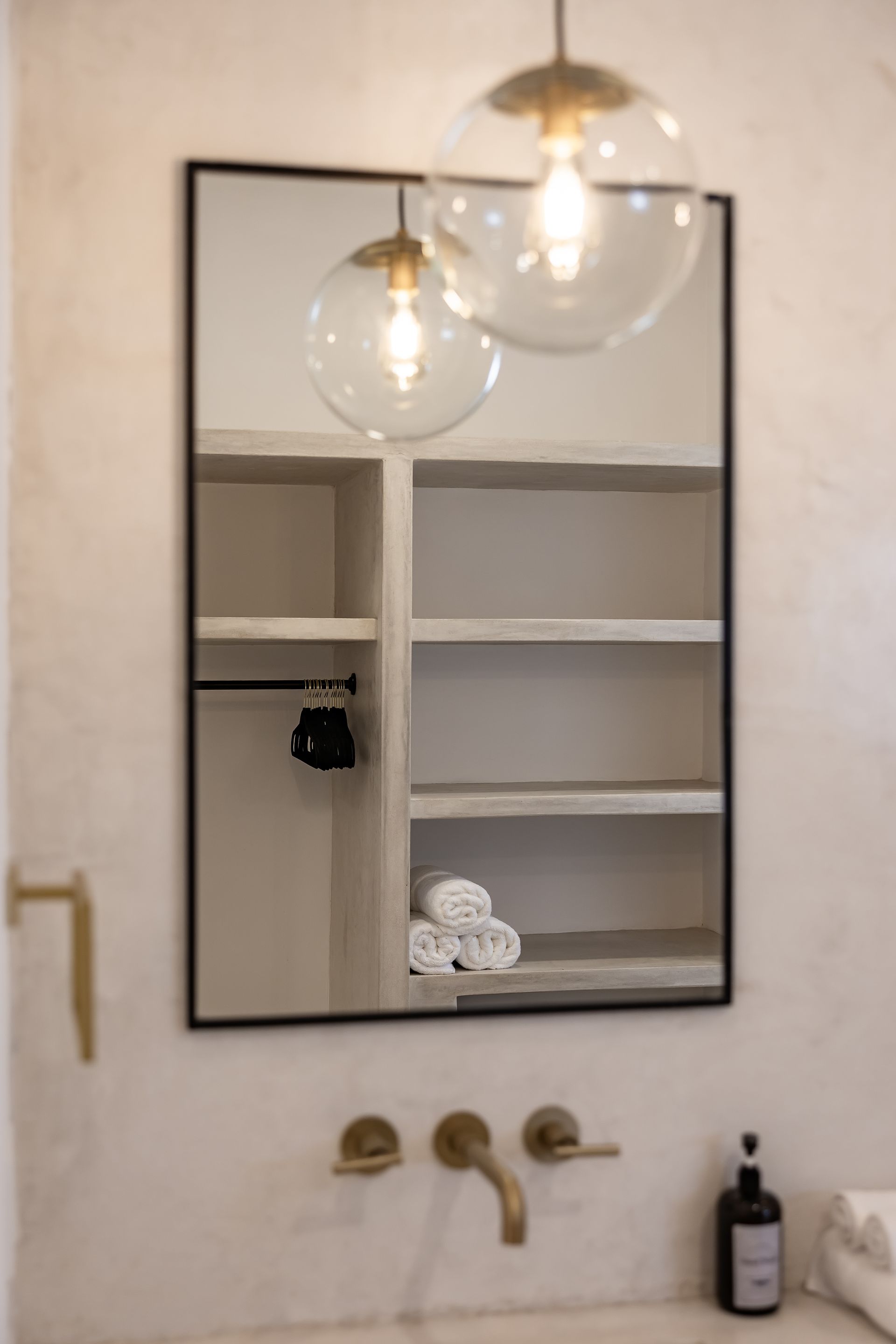Modern bathroom with a black-framed mirror, globe light fixture, wall-mounted brass faucet, and open shelving with towels.