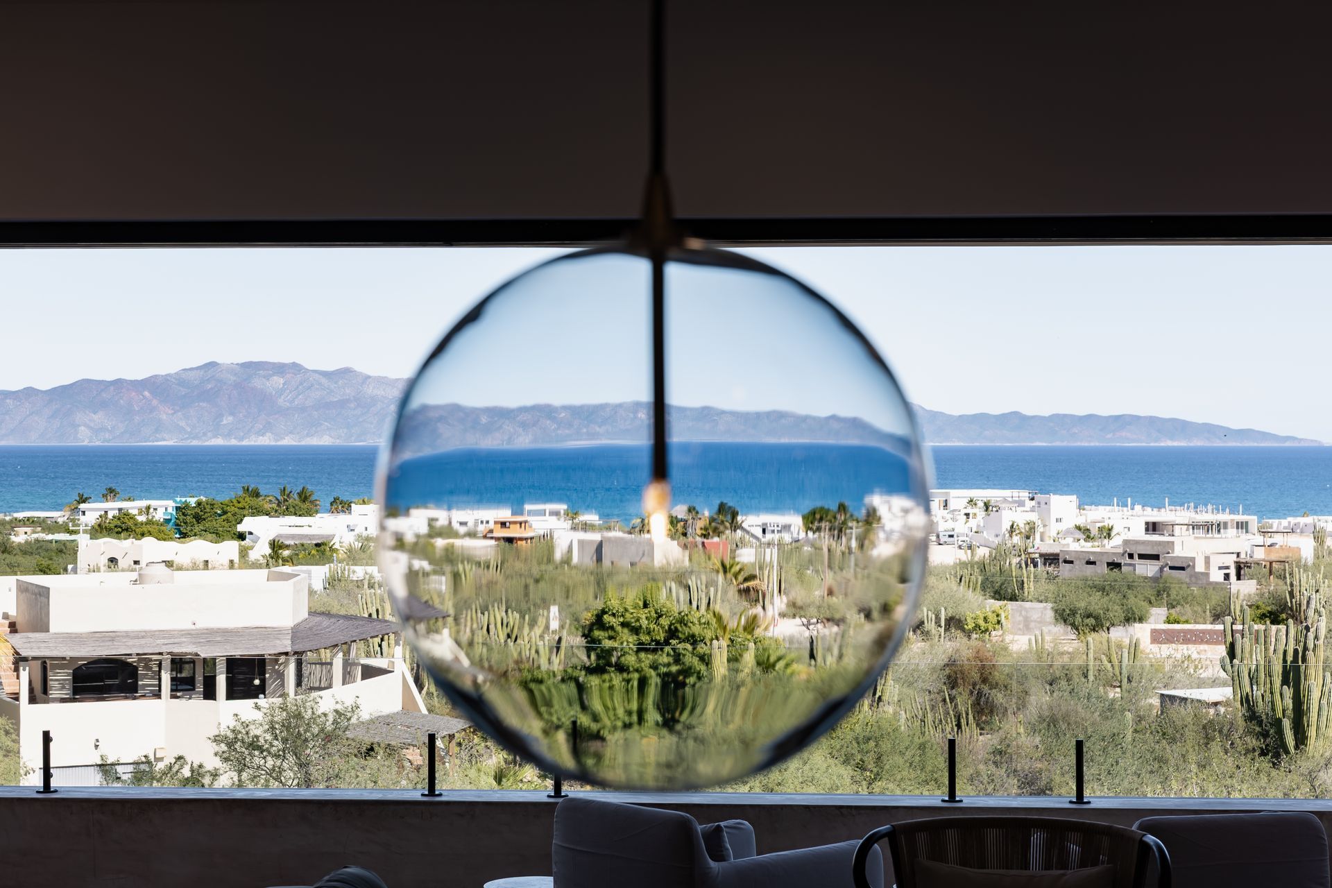 A glass pendant light hangs in front of a window, showing a distorted, inverted view of a coastal town and sea.