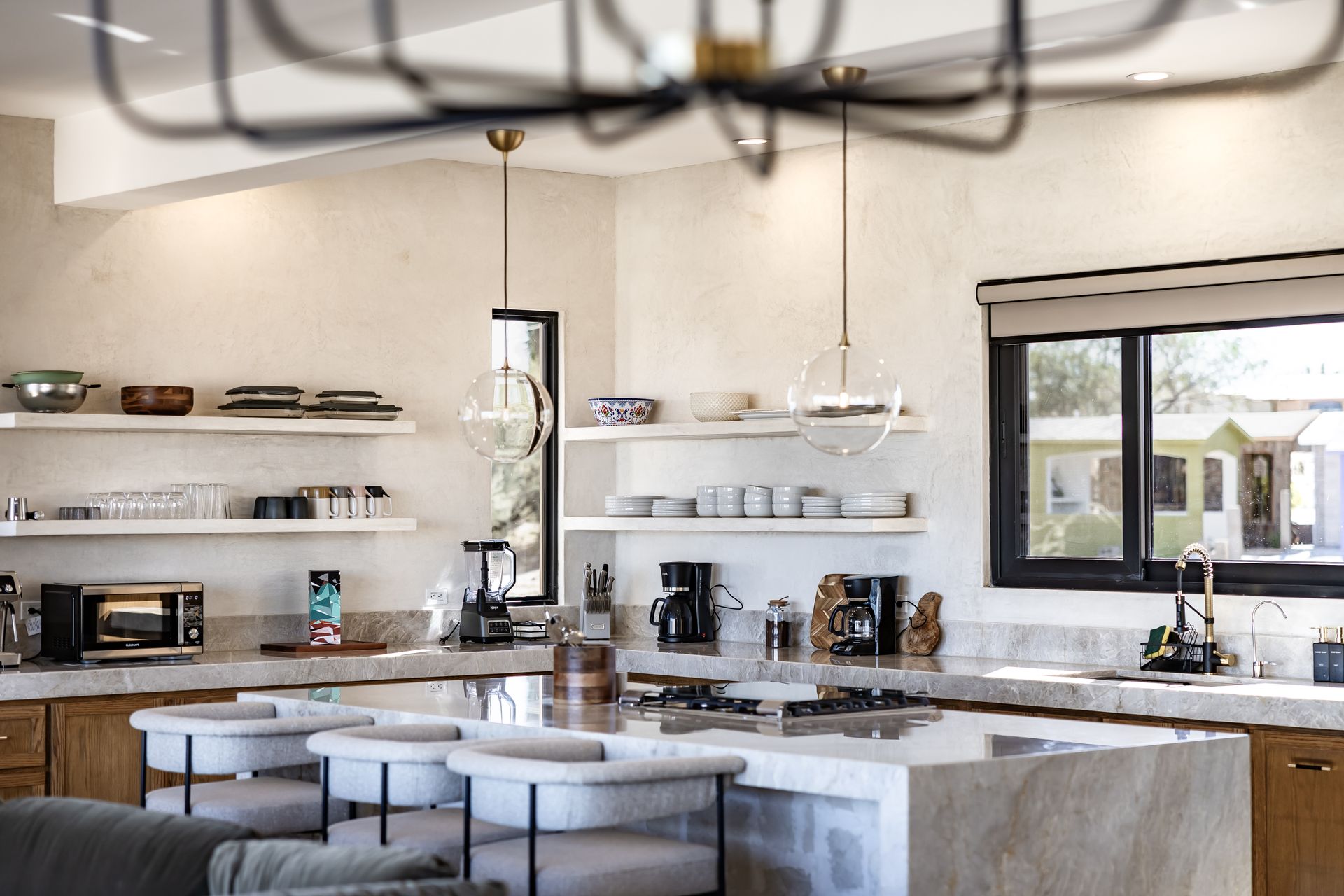 A bright kitchen with marble island, open shelving, and pendant lights, featuring wooden cabinetry and large windows.