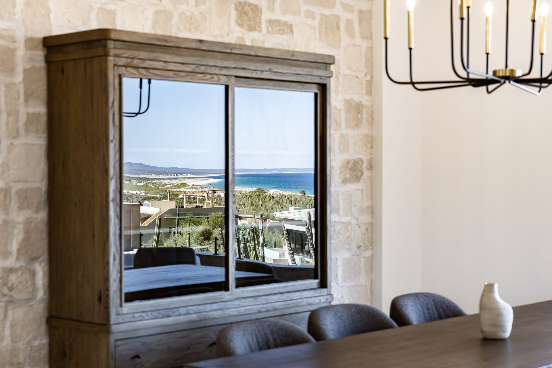 A wood-framed cabinet mirror reflects an ocean view, set against a stone wall next to a dining table and pendant light.