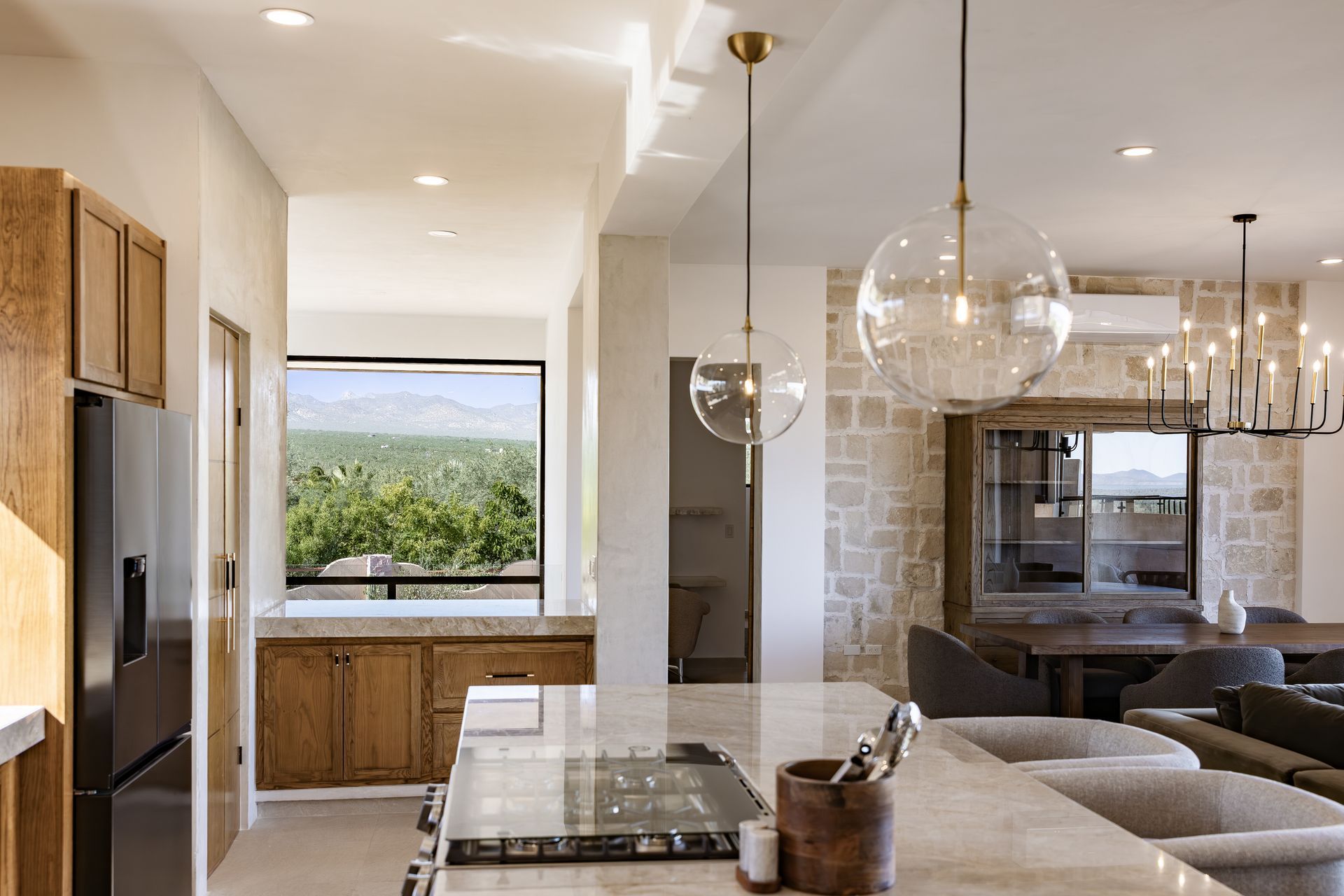 A modern kitchen with wood cabinets, marble counters, glass pendant lights, and a stone wall overlooking a mountain view.