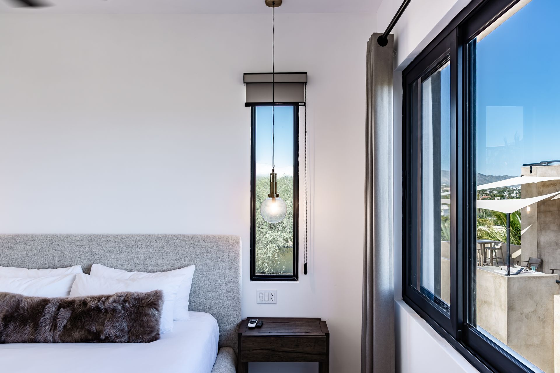 A bedroom featuring a bed with a textured headboard, a narrow vertical window with a pendant light, and a patio view.