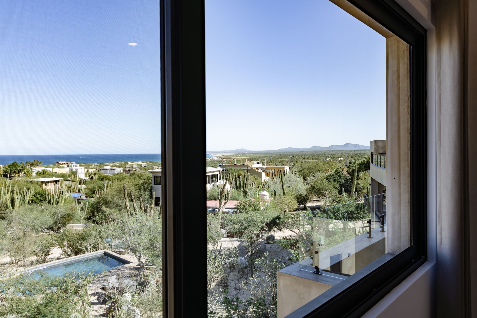A window view showing a sunny coastal landscape with lush green trees, a small pool, and the ocean in the distance.