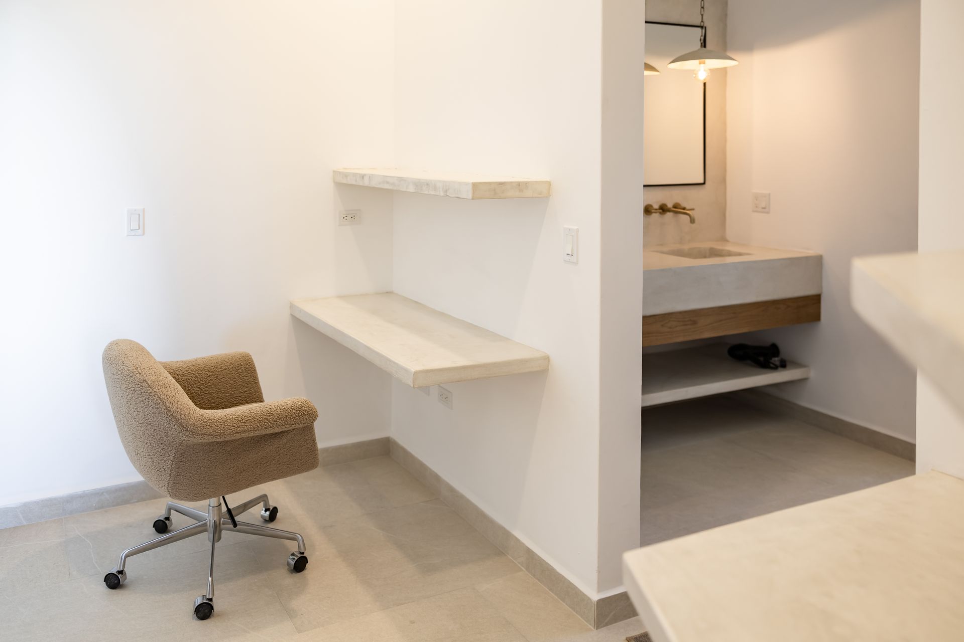 A desk area with a textured armchair and two wall-mounted shelves, adjacent to a modern bathroom with a stone sink.