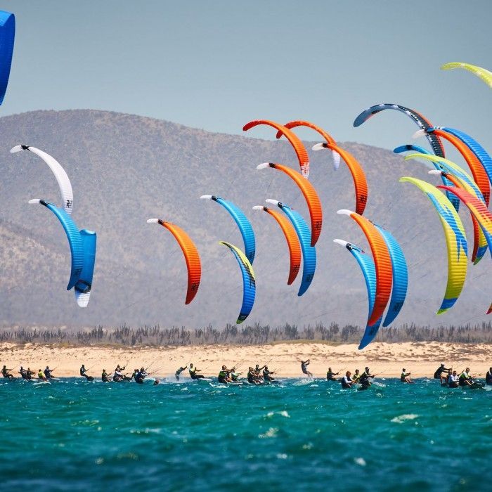 Kite surfers in colorful kites over blue water near a sandy beach. Hills are in the background.