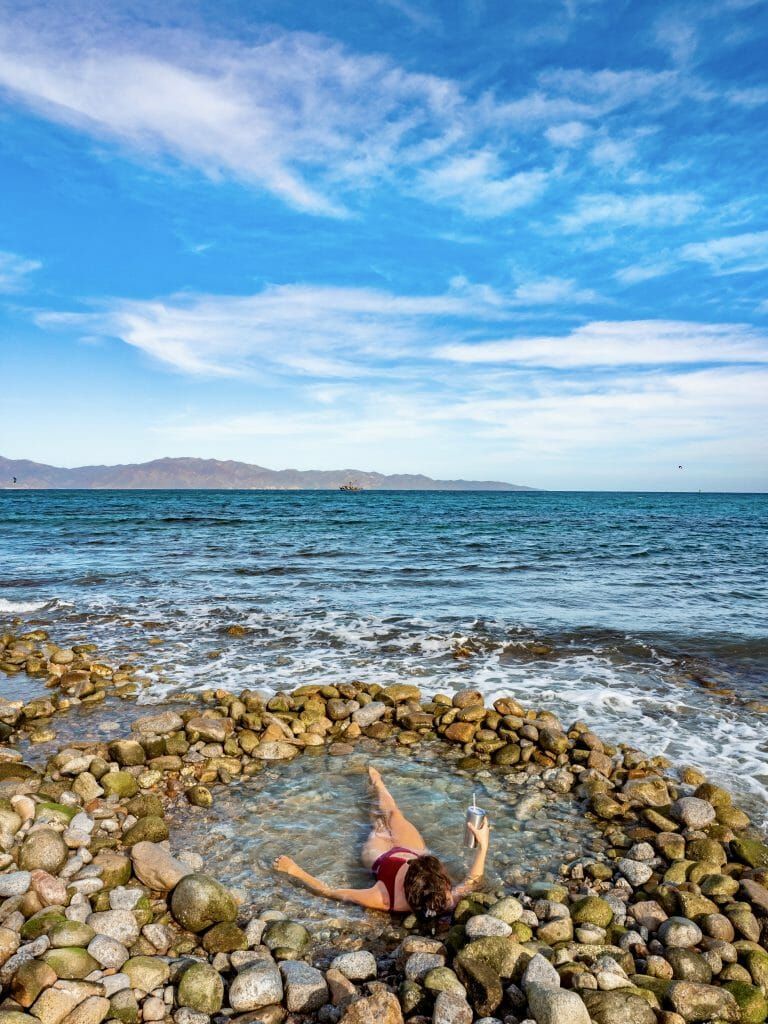 Woman relaxing in a stone-lined pool on a beach. Ocean and blue sky background.