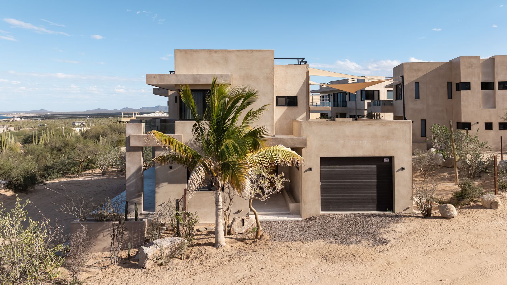 Modern beige multi-story house in a desert landscape with a palm tree, a dark garage door, and a clear blue sky.
