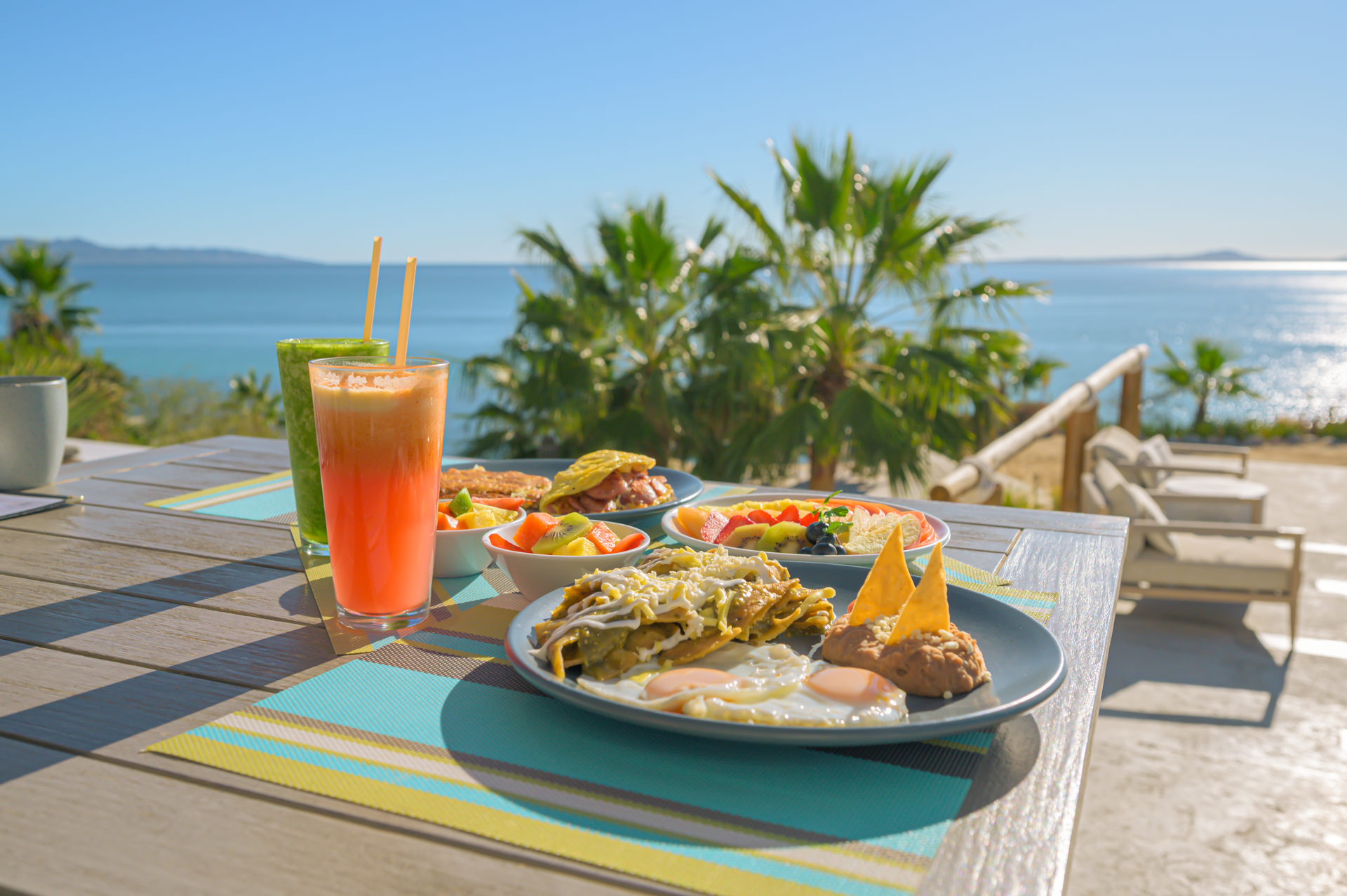 A breakfast spread with juice and chilaquiles on a table overlooking the ocean on a sunny day.