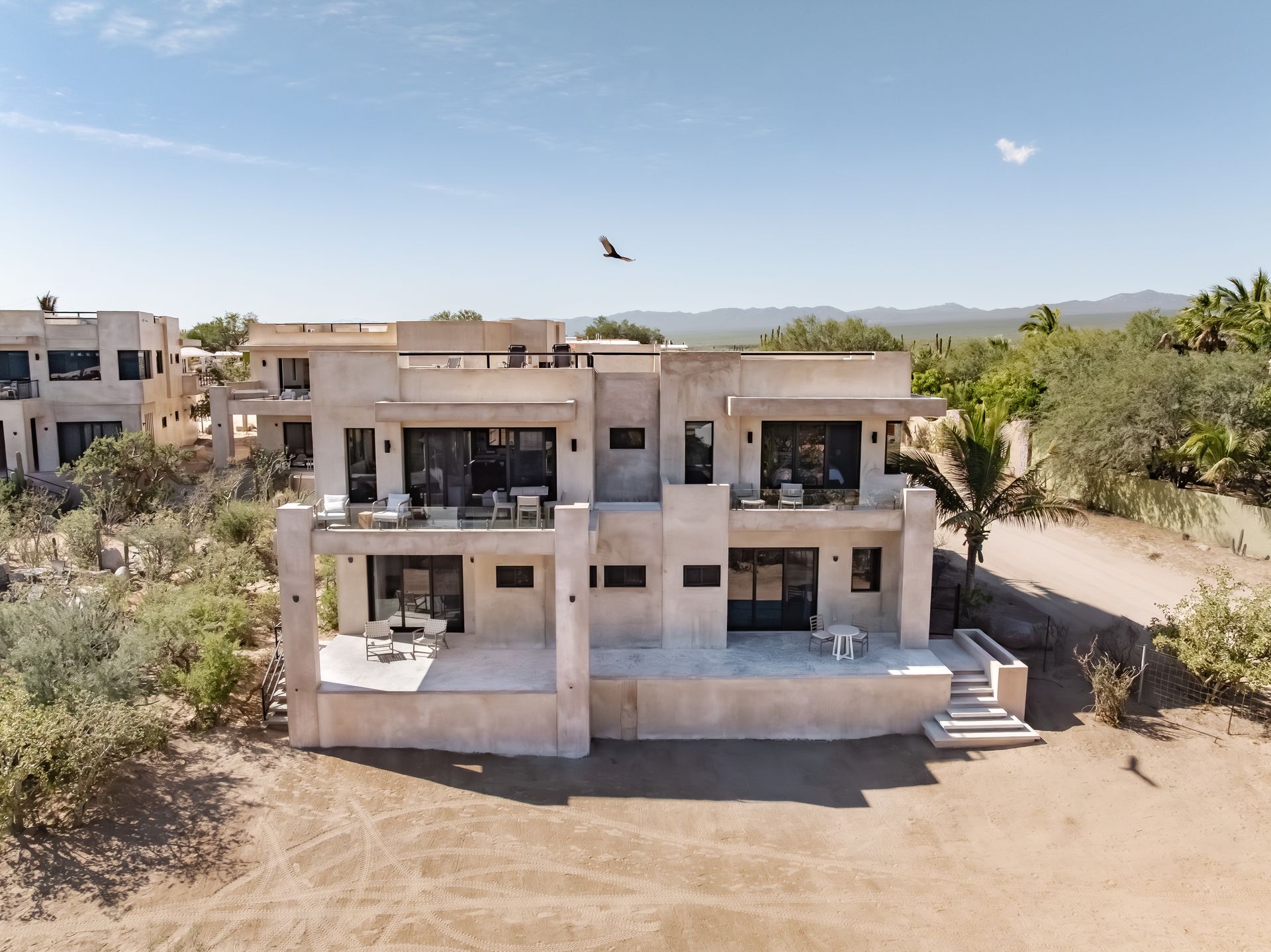 A tan, two-story modern house with flat roofs and balconies, situated in a dry, arid landscape under a clear blue sky.