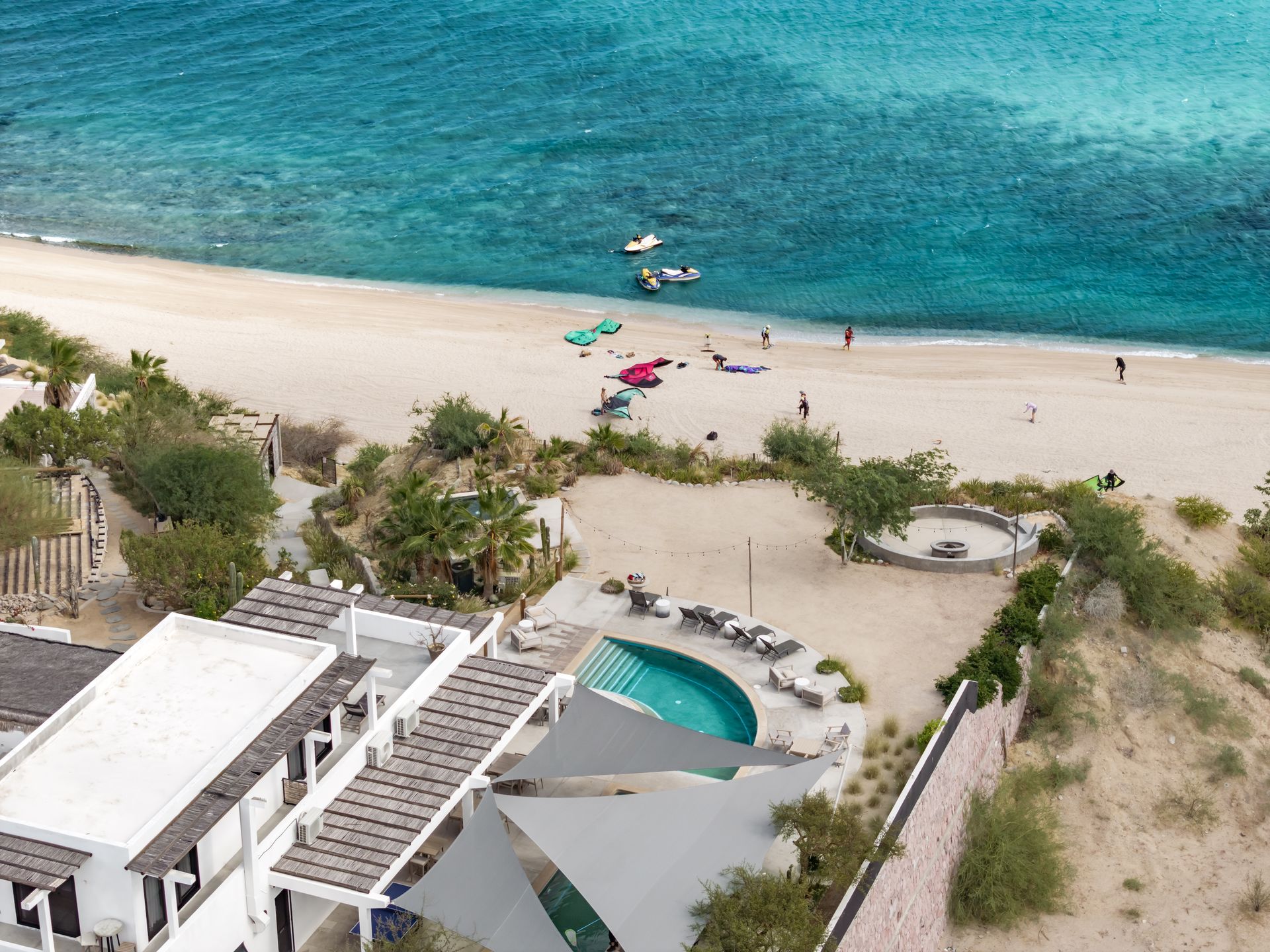 An aerial view shows a white building with a pool and patio beside a sandy beach and turquoise ocean.