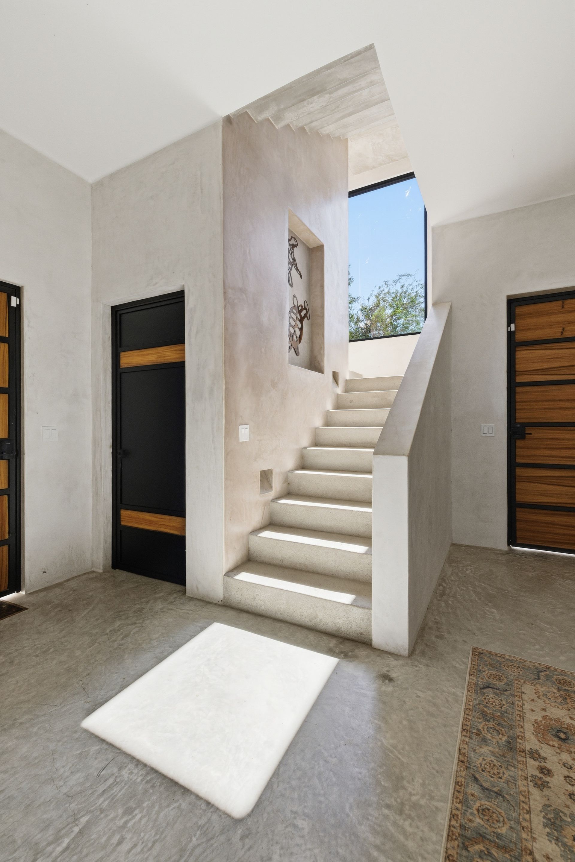A modern interior features concrete stairs leading to a sky-lit landing between two doors with dark, wood-accented panels.