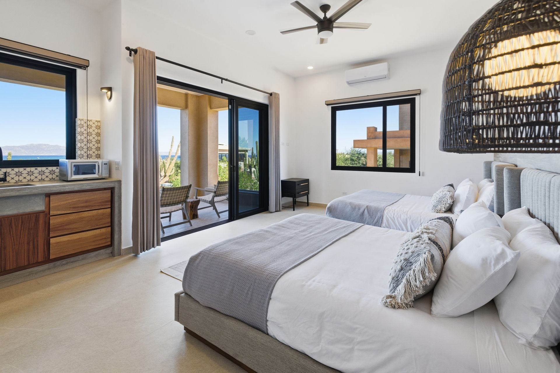 A bright hotel bedroom with two beds, a private balcony, a wooden dresser, and a large woven pendant light.
